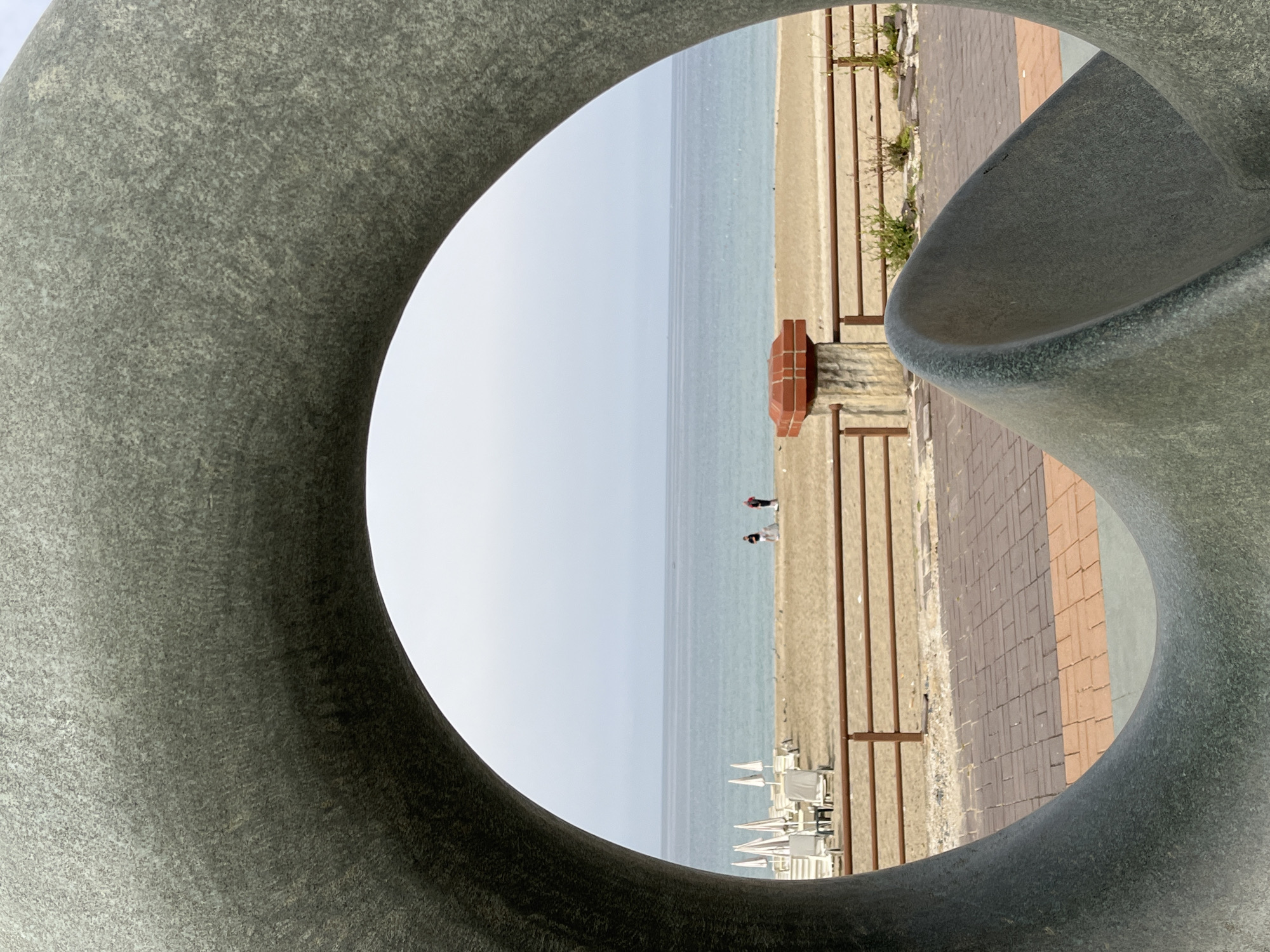 The image shows a view of the Mediterranean Sea and a beach in Alanya, Turkey, framed by a gray concrete sculpture. Two people are walking near the water's edge. White beach chairs are arranged in rows on the sand. A paved walkway runs parallel to the beach, separated by a low fence. A small brick structure is visible on the walkway. The sky is pale blue. The sculpture is composed of twisting, rounded shapes.
