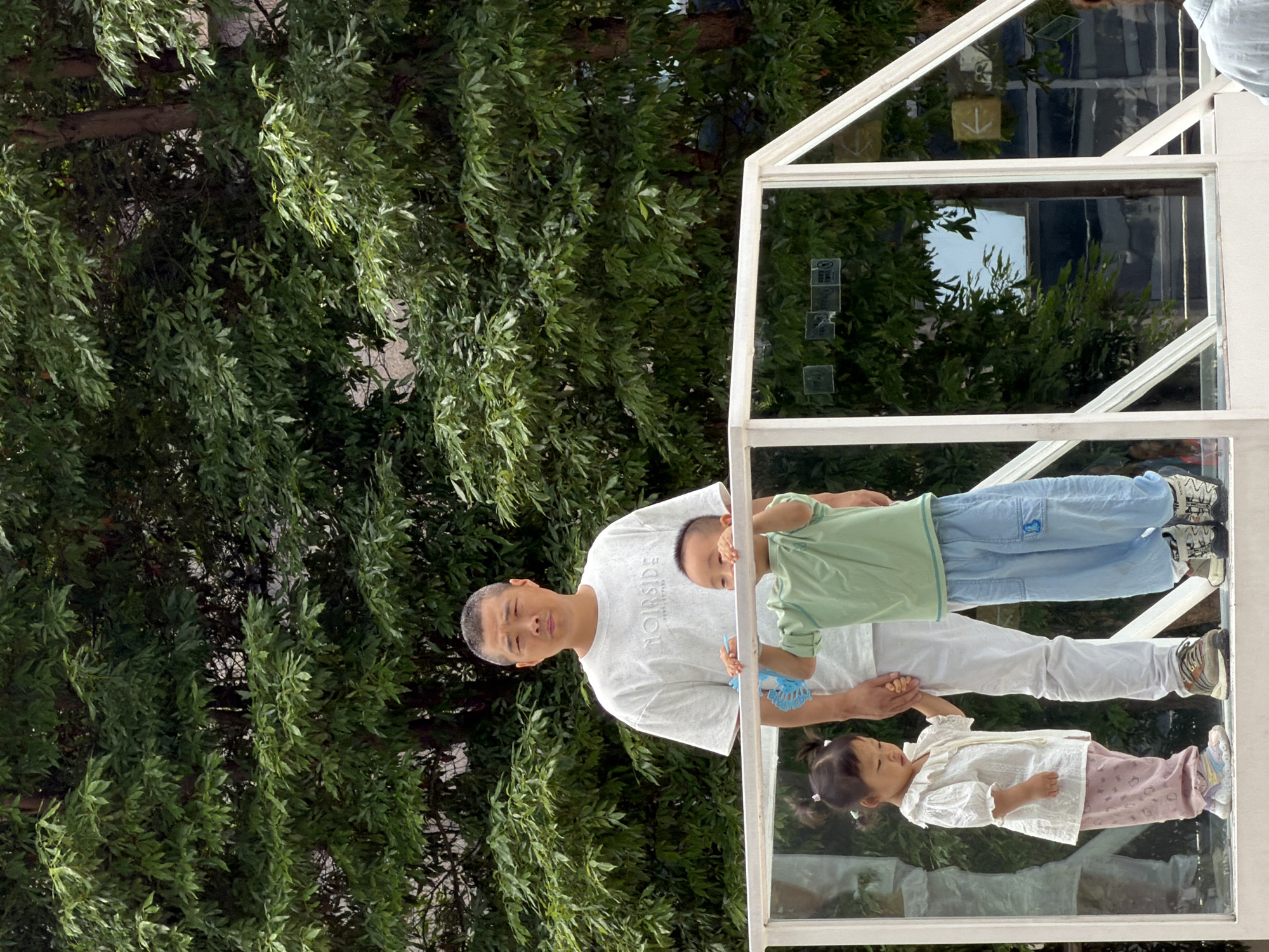 In Yuzhong District, China, a man stands on a white platform behind a glass railing, looking towards the camera with a neutral expression. He is wearing a light gray t-shirt with the text "NOIRSIDE" printed on it and white pants. To his right, a young boy in a light green t-shirt and blue pants stands holding the railing. To the man's left, a young girl wearing a white blouse and pink patterned pants holds the man's hand. Both children are looking away from the camera. Behind them, lush green foliage from trees forms a dense backdrop against a bright sky. The platform features a white railing with diagonal supports, and reflections of trees and buildings are visible in the glass panels of the railing.