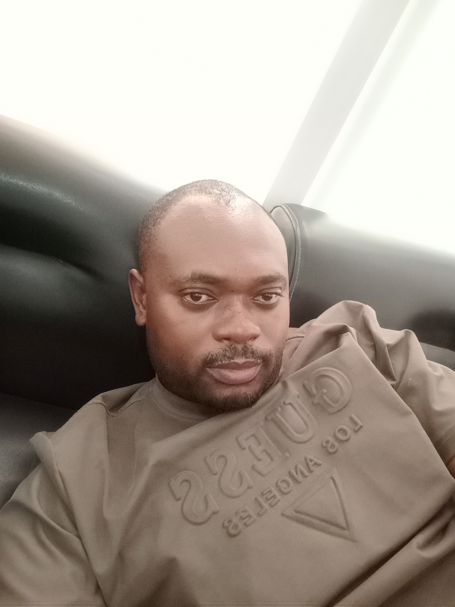 A medium-shot selfie depicts a man with dark skin, short hair, and a groomed beard and mustache. He is wearing a light brown t-shirt with the word "GUESS" and "LOS ANGELES" embossed on it, with the words in reverse writing. The man is sitting on a black leather couch, and a white wall is visible in the background. The location is likely indoors but the context related to the city of Gwarinpa, Nigeria, cannot be verified from the image alone.