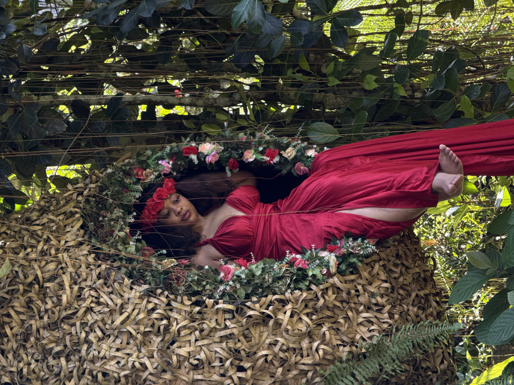 In Tegallalang, Indonesia, a person is seated in an oversized nest-like structure woven from natural, light-colored fibers, adorned with artificial flowers and greenery. The individual, wearing a vibrant red dress with a high slit and a floral headpiece, is positioned within the opening of the nest, with their legs exposed and dangling. The background features dense foliage, including large green leaves and hanging vines, suggesting a lush, natural setting within a tropical environment. Sunlight filters through the leaves, creating dappled light and shadows throughout the scene.