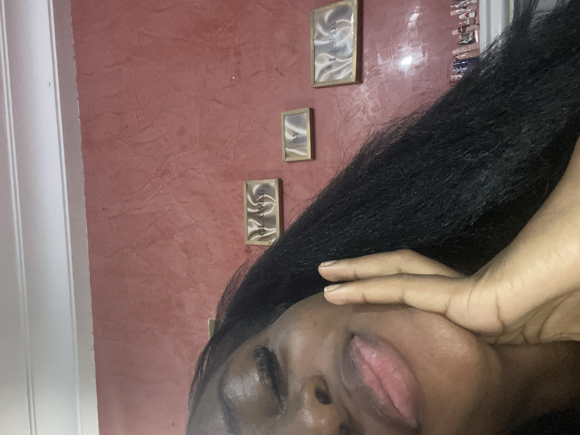 The image shows a close-up of a young Black woman's face, with her right hand resting on her chin and cheek. Her eyes are closed, and her lips are slightly parted, appearing to be coated in a glossy lip product. She has long, dark, wavy hair that falls over her shoulder and partially obscures the background. The background consists of a textured, reddish-pink wall with white molding at the top. Three small, framed pictures are hung on the wall, arranged horizontally. The frames appear to be made of wood. On a shelf below the pictures, a row of nail polish bottles is visible. The lighting suggests an indoor setting, likely a home. There is no information available to determine the location of this scene within the city of Jos, Nigeria.