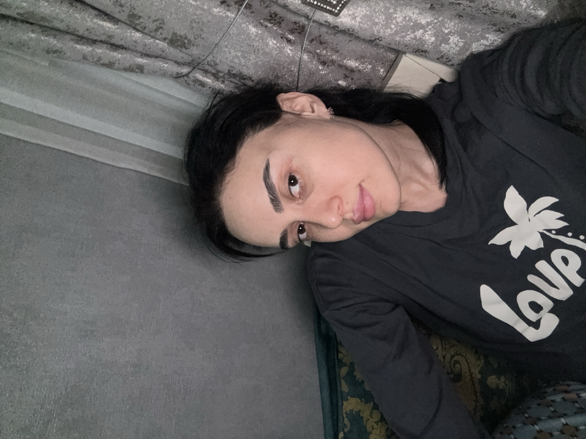 A dark-haired individual is captured in a close-up, indoor shot, likely a selfie, as they lie down. Their head is positioned to the right of the frame, looking towards the camera with a neutral expression, hinting at a slight smirk. They have prominent dark eyebrows and dark eyes. A small, light-colored earring is visible in their right ear. The person is wearing a dark, long-sleeved shirt featuring a white graphic design. The word "Love" is clearly visible in white, styled with a white palm leaf or tropical floral icon above and to the left of the text.
The setting appears to be an interior space, possibly a living room or bedroom, indicated by the soft furnishings. Behind the person's head, a textured, grey and white patterned curtain or wall covering is visible. To the left, a plain grey wall or surface is present. Below the person's right arm, there is a glimpse of a dark green fabric with an ornate gold pattern, suggesting a blanket, cushion, or upholstered furniture. The lighting is artificial, characteristic of an indoor environment, making the time of day undeterminable. No explicit cues in the image indicate the location as Tbilisi, Georgia, but this is provided as context.