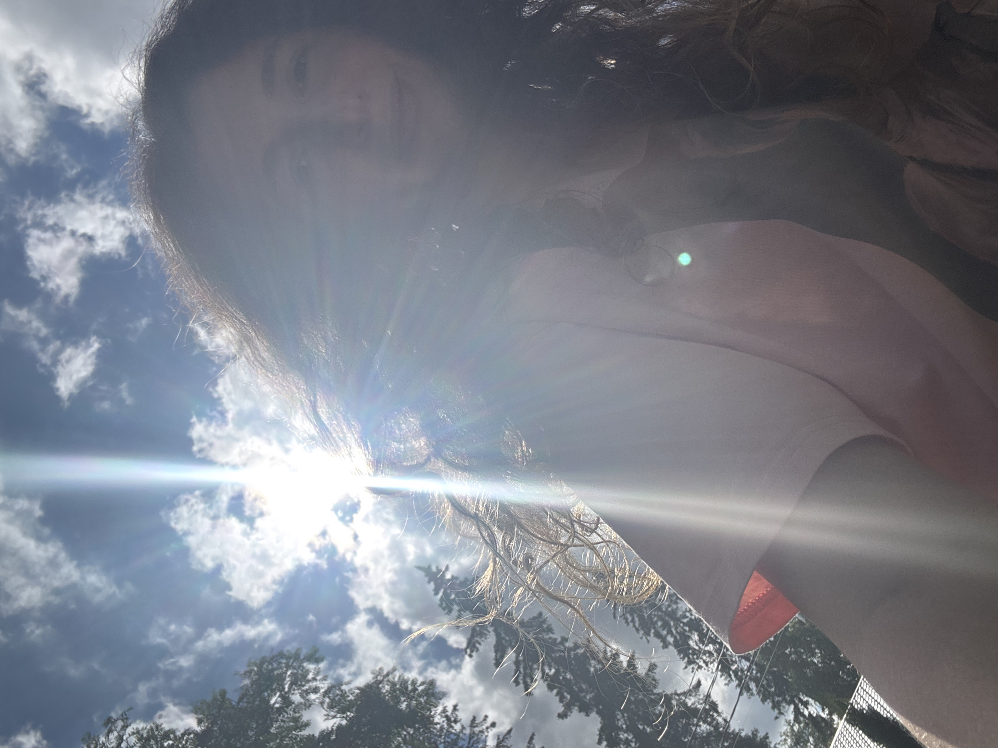 An outdoor, low-angle, selfie-style photograph shows a person with long, dark, curly hair and fair skin in the foreground. The person is wearing a light pink t-shirt. The sun is bright and positioned in the upper center of the frame, causing lens flare and partially obscuring the person's face. The background consists of a blue sky with scattered white clouds and the silhouettes of trees on the left and right sides. A thin, white fence is visible at the bottom left of the frame. The image appears to have been taken during the daytime.