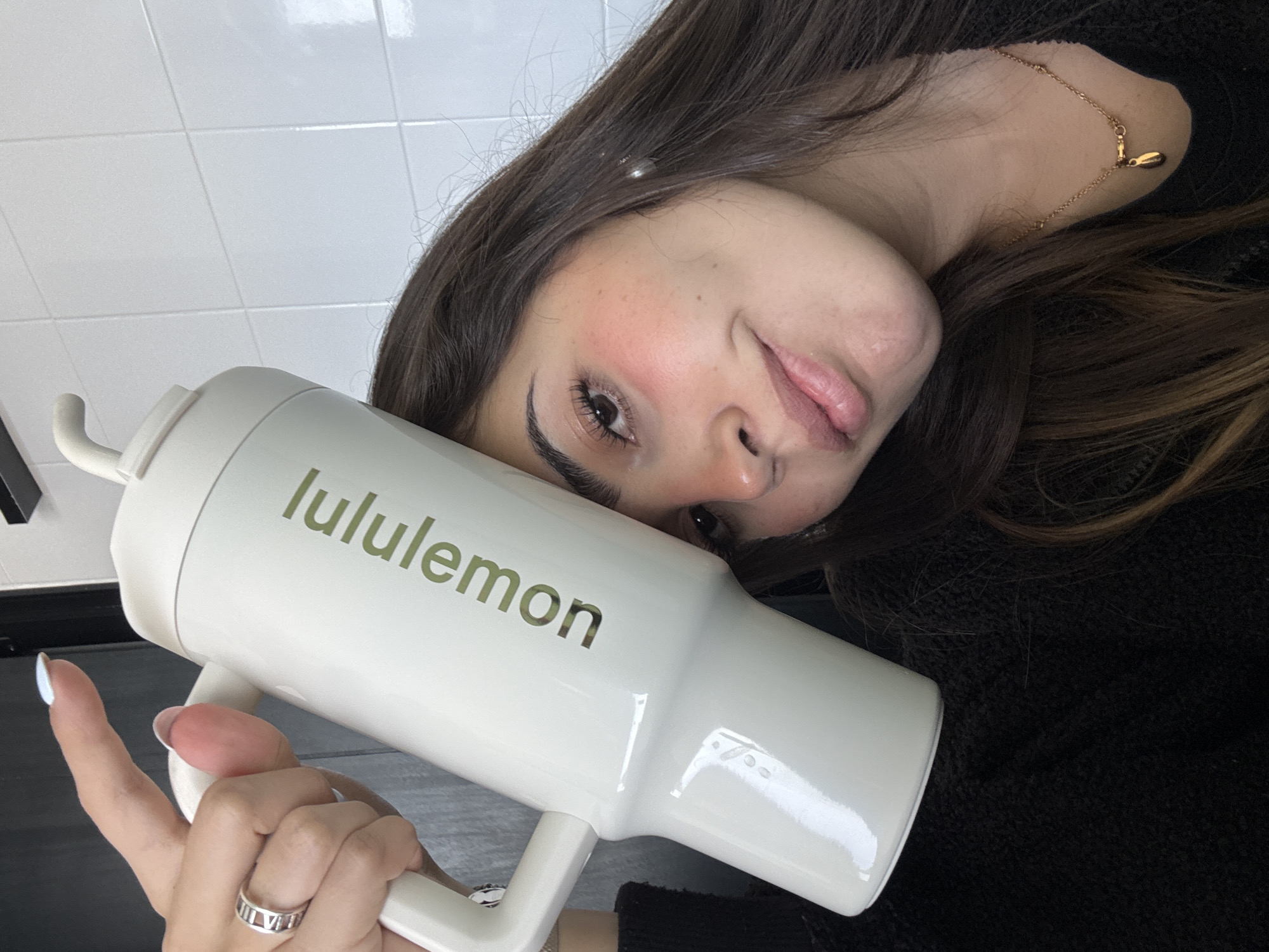 A young woman is captured in a close-up, overhead portrait, possibly a selfie, set indoors against a backdrop of white tiled walls, likely in San Bernardino Tlaxcalancingo, Mexico. Her long, dark brown hair cascades around her head and shoulders, and she is wearing a dark, textured top. She holds a large, light-colored, handled tumbler directly in front of her, angled towards the camera. The word "lululemon" is prominently displayed in a greenish-gold font on the side of the tumbler.
The woman's head is tilted, and she looks towards the camera with a subtle, perhaps playful, expression, her lips slightly parted in what appears to be a hint of a smile or pout. Her makeup includes noticeable pink blush on her cheeks and defined eyebrows and eyelashes. She wears a delicate, gold-colored chain necklace with a small, tear-drop shaped pendant visible on her left shoulder. On her left ring finger, a silver-colored ring featuring Roman numerals is visible. Her fingernails are painted with a light-colored polish. The overall mood of the image is casual and direct.