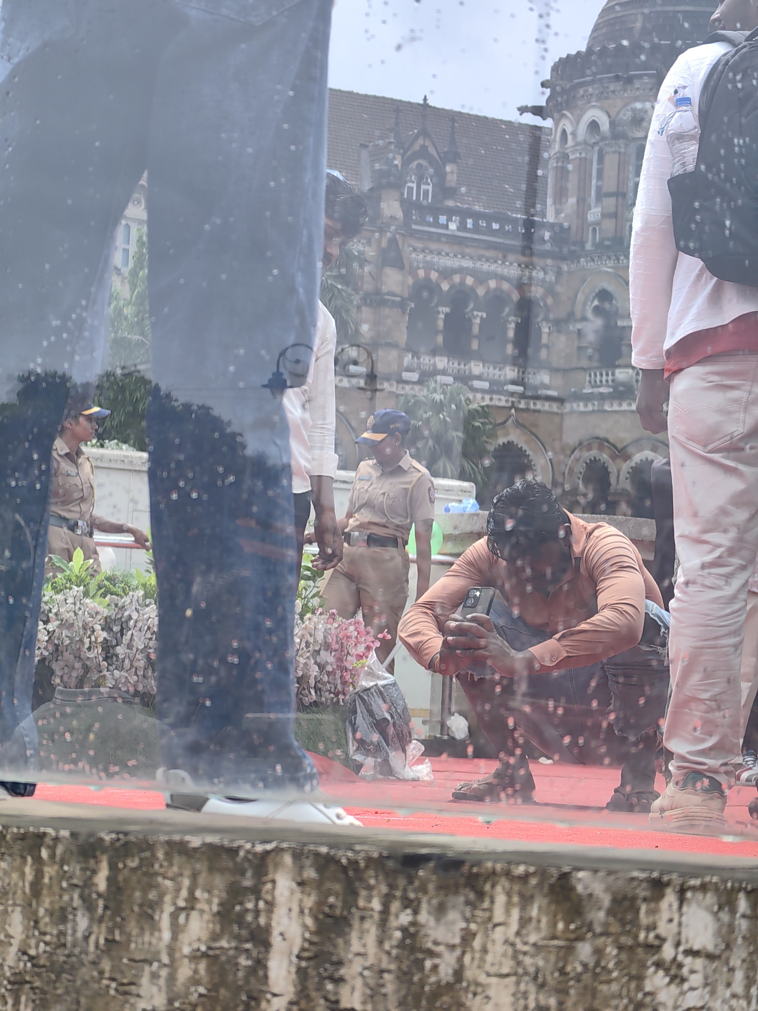 The image depicts a scene outside a grand, historic building, likely CSMT (Chhatrapati Shivaji Maharaj Terminus) in Mumbai, India, due to its distinctive Victorian Gothic architecture with domes, arches, and ornate stonework. The foreground is partially obscured by a reflective surface, possibly a window, with numerous water droplets or condensation.
In the mid-ground, a man in an orange shirt and blue jeans is crouched down, looking intently at his smartphone, which he holds with both hands. He appears to be either taking a photo or recording a video. To his right, the lower legs and feet of a person wearing light-colored pants and sneakers are visible.
Further back, two individuals in police uniforms are present. One, a woman with a blue cap, stands to the left, looking towards the right. The other, a man also wearing a blue cap, is positioned slightly further back and to the right. They are both wearing khaki-colored uniforms.
The ground is covered with a vibrant red carpet, contrasting with the weathered stone of the building. Some potted plants with pink and white flowers are visible behind the uniformed individuals. The overall atmosphere suggests a public space, possibly during an event or a busy day at the landmark station. The presence of police officers implies security measures or their regular duty at this location.