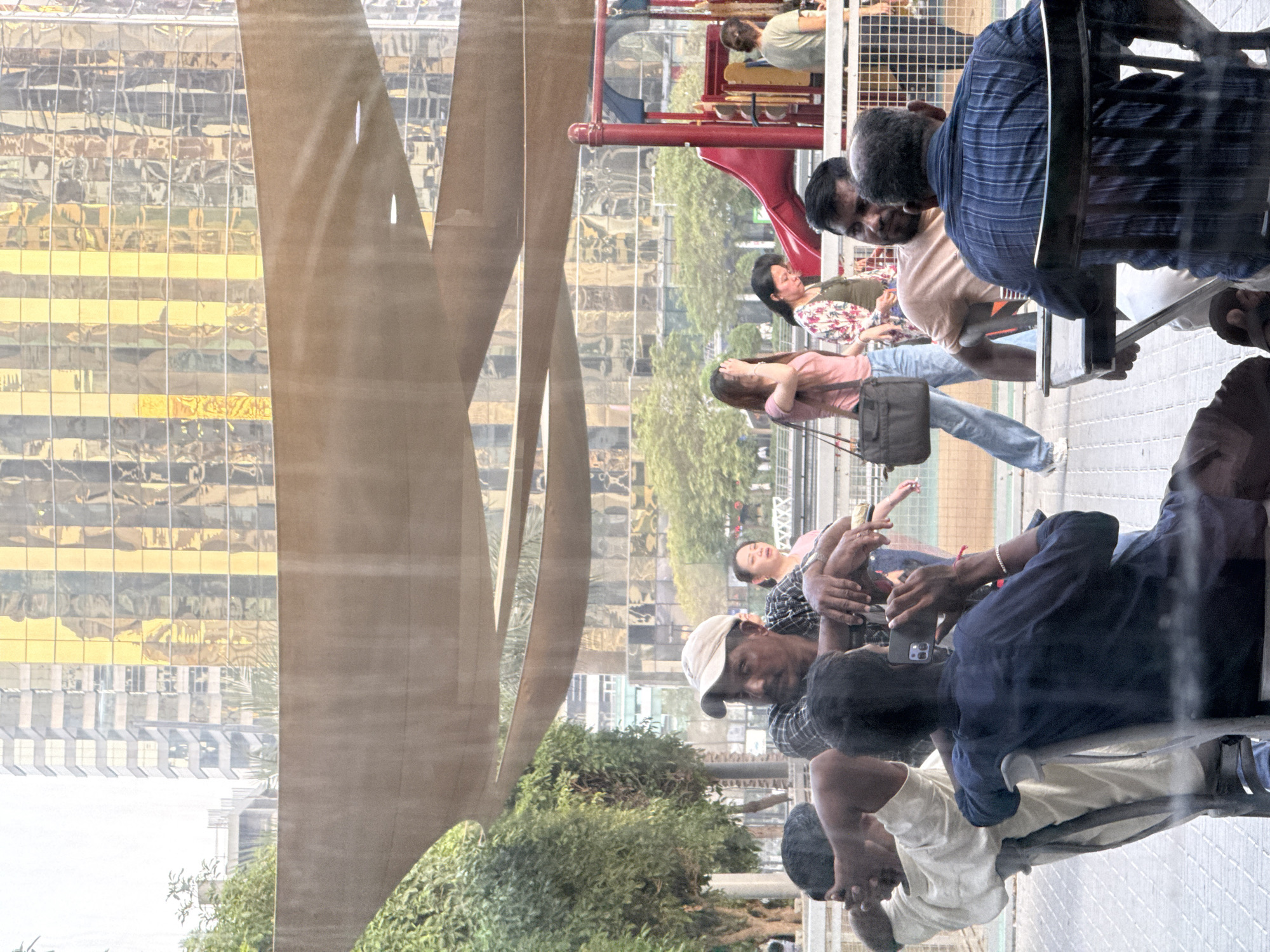 The image depicts an outdoor seating area in Dubai, United Arab Emirates, with a modern cityscape in the background. The foreground features several individuals seated at tables and chairs, some interacting with each other. One man wears a plaid shirt and a cap, leaning forward. Another person in a blue shirt is holding a smartphone. Behind them, several women are visible, some walking and others standing. In the background, a playground with red and blue structures is present, set against the backdrop of a tall building with a reflective, gold-tinted facade, characteristic of Dubai's contemporary architecture. The overall atmosphere appears to be a casual gathering in a public urban space.