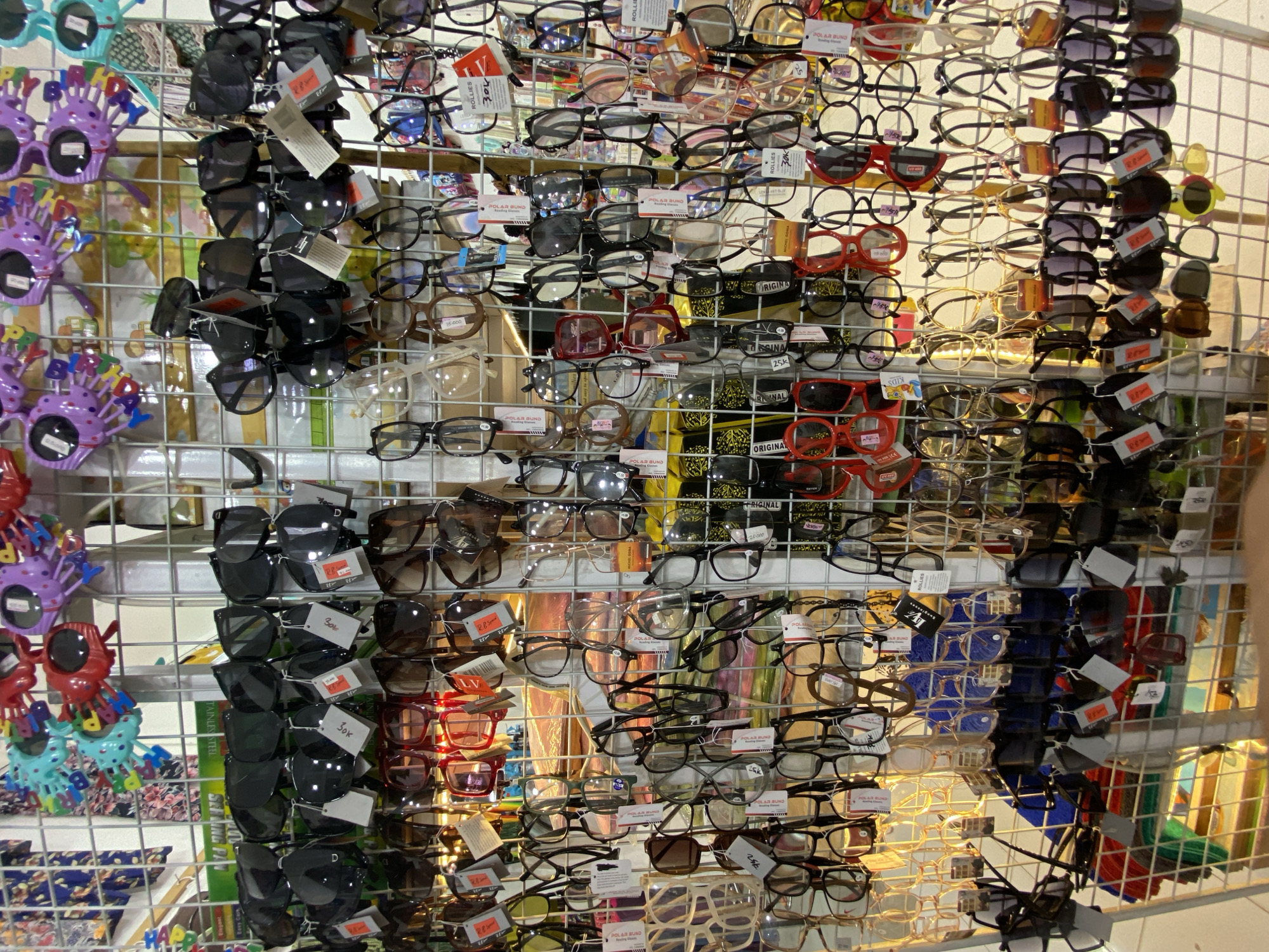 This media file showcases a vibrant display of eyewear, likely sunglasses and eyeglasses, organized on metal racks. The items are varied in style and color, with many pairs featuring dark lenses. Some glasses are attached to colorful, novelty party glasses, such as purple ones with designs resembling flowers or a character's head, and red ones with decorative elements. There are also individual pairs of glasses in various frames, including black, clear, and patterned. Several price tags and labels with text such as "ROLLIES," "POLAR BUND," "ORIGINAL," and "KIDS" are visible, along with numerical price indications like "30k" and "25k." The background suggests an indoor market or store setting, with hints of colorful fabrics or displays behind the eyewear. The lighting appears to be artificial, casting a warm glow on the merchandise, suggesting it might be daytime or evening within a well-lit retail space. The overall impression is one of a bustling point of sale, with a wide assortment of affordable eyewear available to customers. The location context provided is Lamteumen Barat, Indonesia.