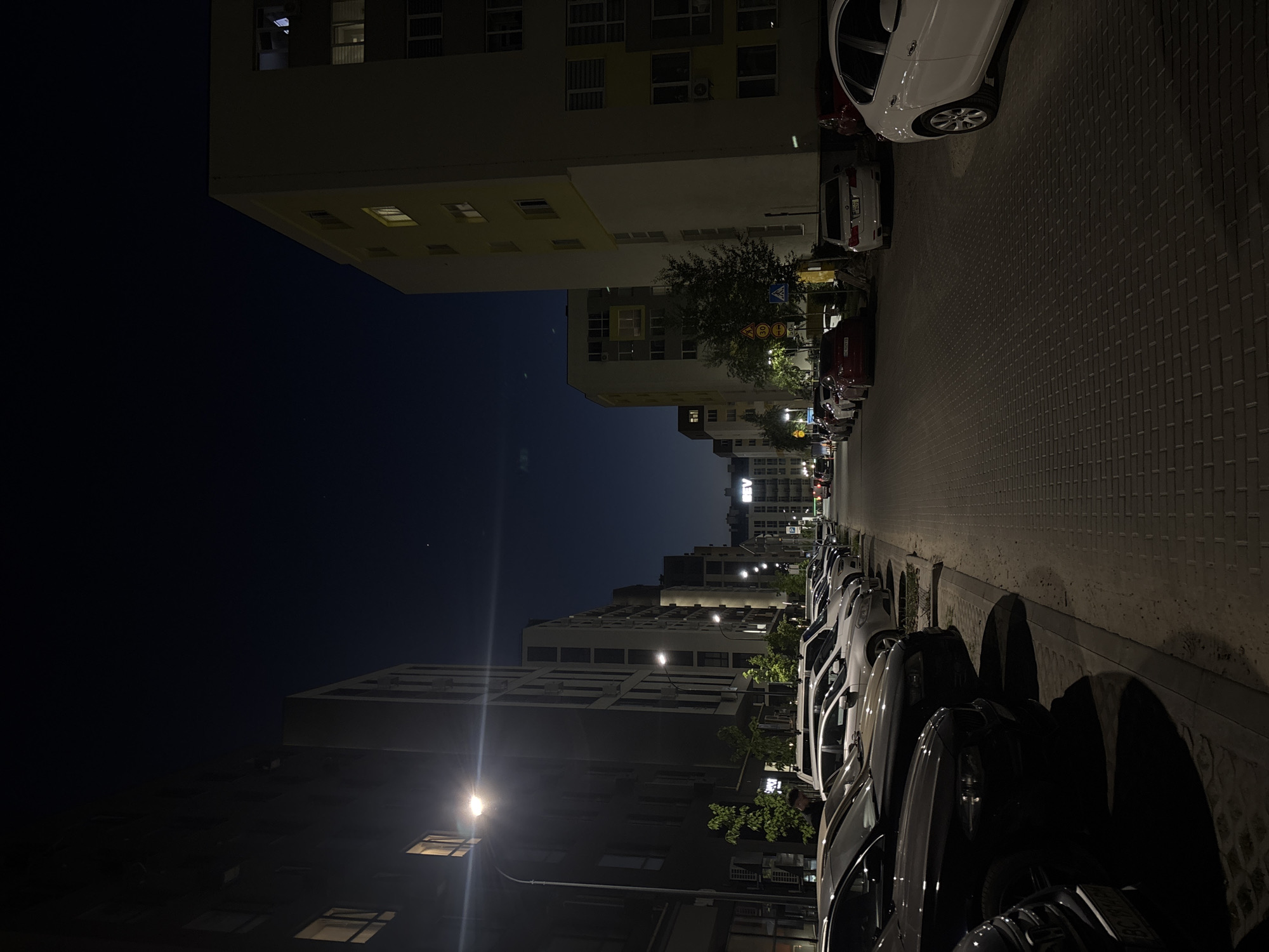 The photograph depicts an urban street scene at night in Sokilnyky, Ukraine. The street is lined with parked cars on the right side, and buildings rise on both sides. The sky is dark, suggesting it is late evening or night. Streetlights illuminate the scene, casting shadows along the road and highlighting the facades of the buildings. The road is paved with bricks. There is no apparent activity, interaction, or people.