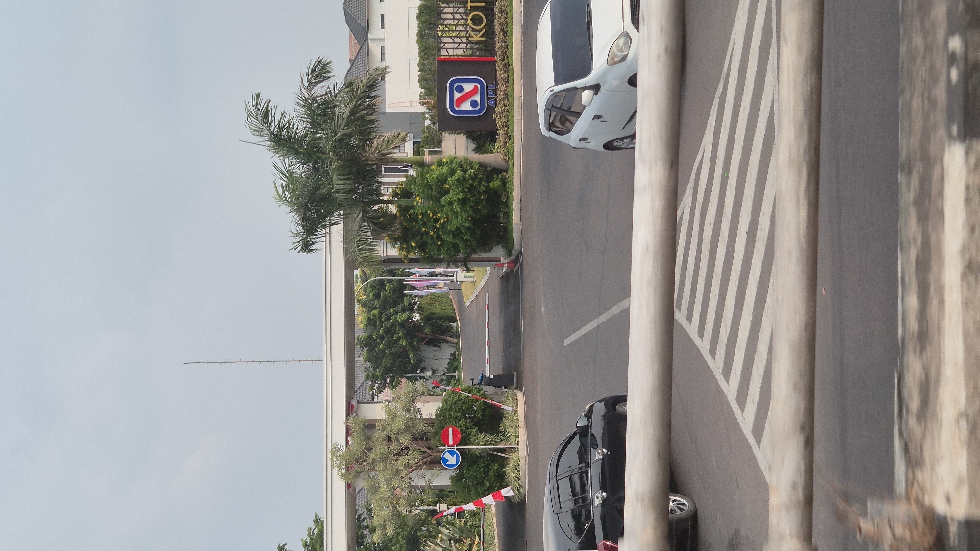 The image depicts the entrance to a residential complex in Karawang, Indonesia. Two cars are visible on the road: a black sedan on the left and a white hatchback on the right. The entrance features a white archway with a barrier gate. To the left of the entrance, there are traffic signs indicating a left turn and a no-entry zone. A billboard for "APL KOT..." is prominently displayed on the right side of the entrance, with a stylized logo. Palm trees and other greenery surround the entrance, and several flags are planted along the road leading into the complex. The sky is overcast. The foreground is partially obscured by a wooden railing and a pedestrian crossing marked with diagonal stripes.