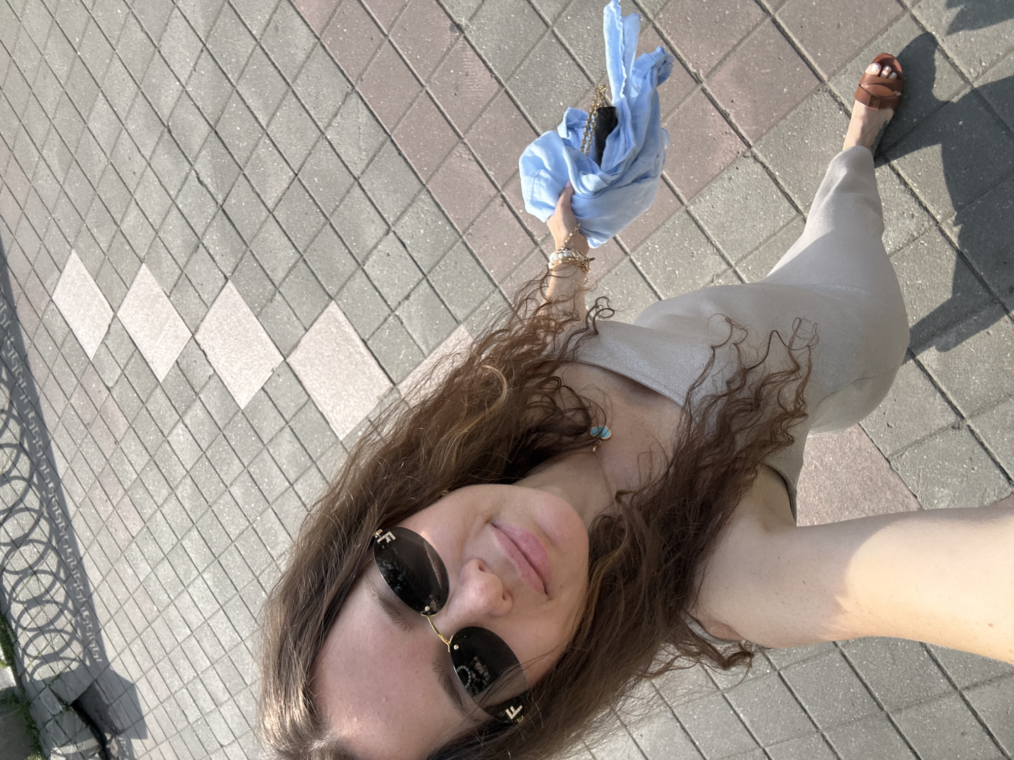 A young woman stands on a paved sidewalk composed of square and rectangular gray and pinkish-red tiles. She is wearing a light-colored, form-fitting dress, brown sandals, and dark-rimmed sunglasses with gold accents. She holds a crumpled light blue garment in her right hand, which also contains a small black purse with a gold chain strap. The woman has long, wavy brown hair and is looking towards the camera with a slight smile. A shadow from a decorative metal fence is cast on the sidewalk to the upper left of the image. The environment appears to be an outdoor urban setting, likely a street or park area in Yekaterinburg, Russia, given the architectural style of the fence casting a shadow and the common use of this type of paving stone. The lighting suggests it is daytime, with the sun casting distinct shadows.
