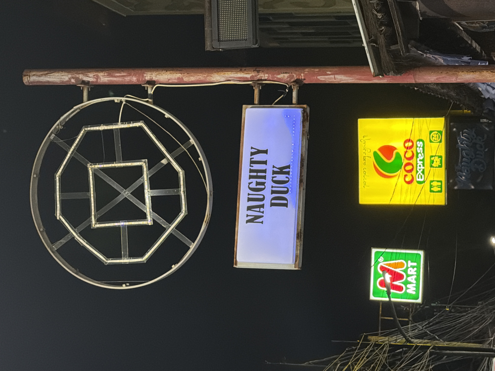 The image shows several signs illuminated at night. The sign reading "NAUGHTY DUCK" is a vertical rectangle with blue illumination. To the left is a round sign, framed with light strips in a geometric pattern. To the right is a yellow sign advertising "COCO Express". Below the "COCO Express" sign, there's a sign displaying "MART" in green and red. The signs appear to be hanging from a rusty, horizontal bar. The location is Kuta, Indonesia, given the presence of the "NAUGHTY DUCK" sign. Below the signs, there are numerous electrical wires and cables.