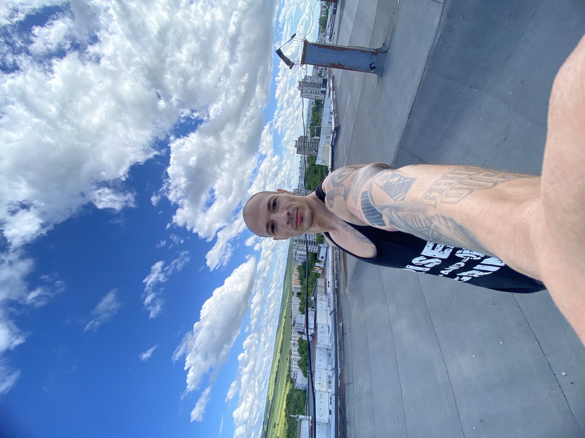 A bald man, appearing to take a selfie, is the primary subject of this image. He is wearing a black tank top, and his arms, visible in the foreground, are heavily tattooed with intricate designs and some text. His expression is neutral to slightly smiling.
The man is positioned on a flat rooftop, which is visible as a grey surface to his right and below him. The scene provides an expansive view of a city or town, identified as Pleven, Bulgaria, with numerous buildings extending into the distance. Beyond the urban sprawl, green fields are visible on the horizon, suggesting a rural transition.
The activity is a selfie being taken from a high vantage point, possibly a building roof. The image is oriented sideways, with the man's head on the left and his body extending towards the right.
The time of day is daytime, indicated by the bright, clear lighting. The weather is sunny with a vibrant blue sky filled with numerous white, fluffy cumulus clouds.
Visible text includes partial words on the man's black tank top, appearing to be in a bold, white font, possibly starting with "STOP" or a similar word, though it's largely obscured. Some faint text is also visible within the tattoos on his left arm, where "FIGHT" might be discernible.