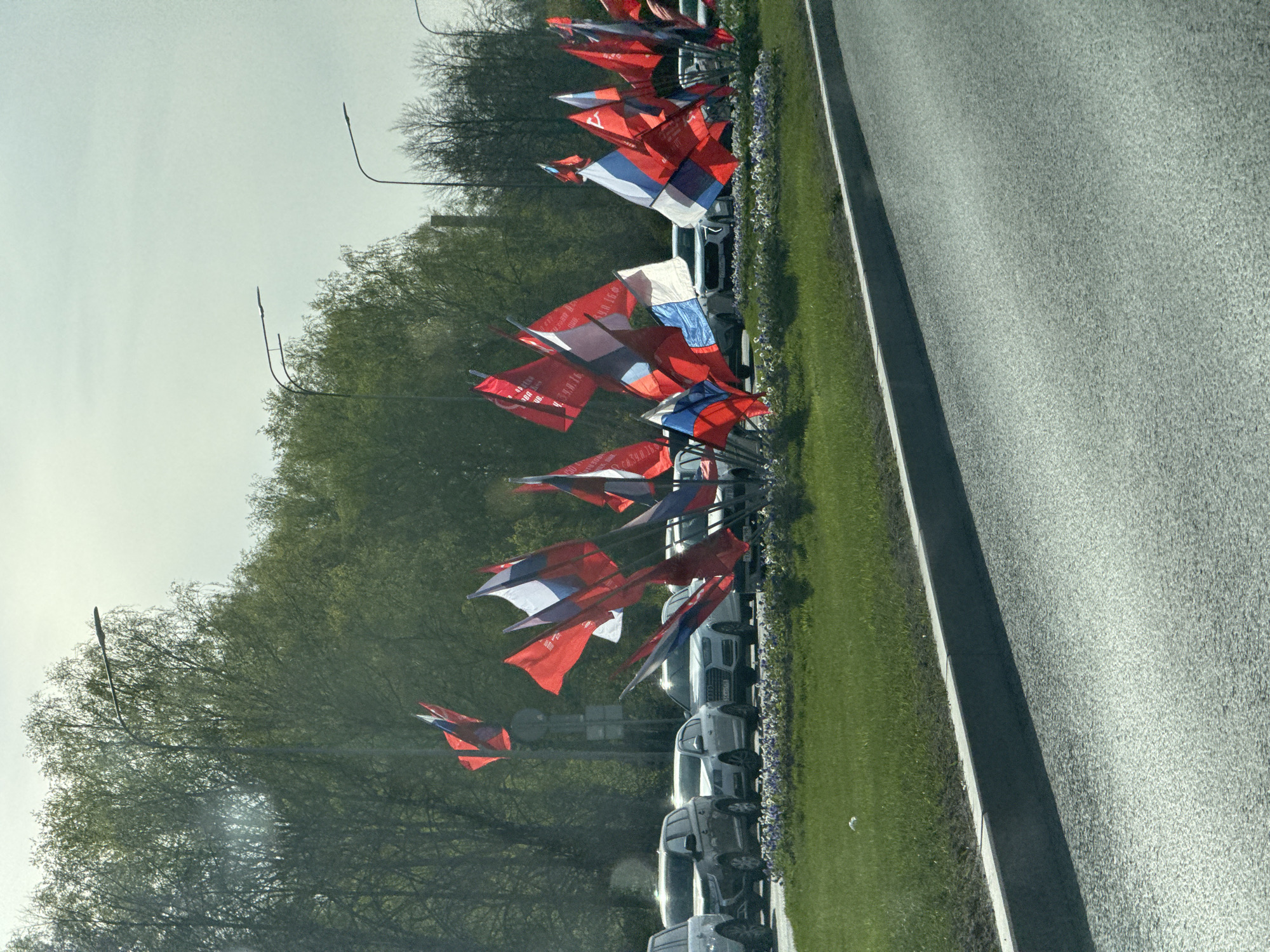 The image shows a row of cars parked along a grassy median strip next to a roadway. Decorative flags, primarily red, white, and blue, adorn the median strip and likely commemorate a patriotic event. Trees line the backdrop, suggesting a parkway or landscaped area. The flags' colors and design suggest Russian national symbols. Based on the prevalence of these flags and the setting, the location is likely in Saint Petersburg, Russia.