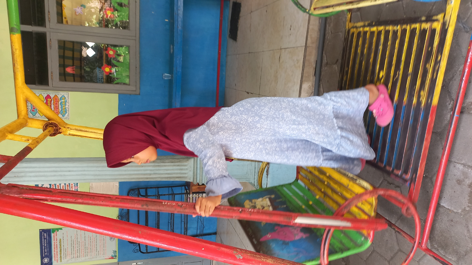 A young girl wearing a maroon hijab and a light blue patterned dress is standing on a playground swing set, holding onto the red metal support pole. She is looking to her left and has her right foot on a yellow and blue striped platform. The swing seat is green and yellow with some pink images on it. The background includes a blue wall, windows with decorations on them, and a yellow metal structure. There are also signs with text visible on the wall to the left. The playground appears to be outdoors, with a paved ground surface. The scene is likely in Pare, Indonesia, given the context.