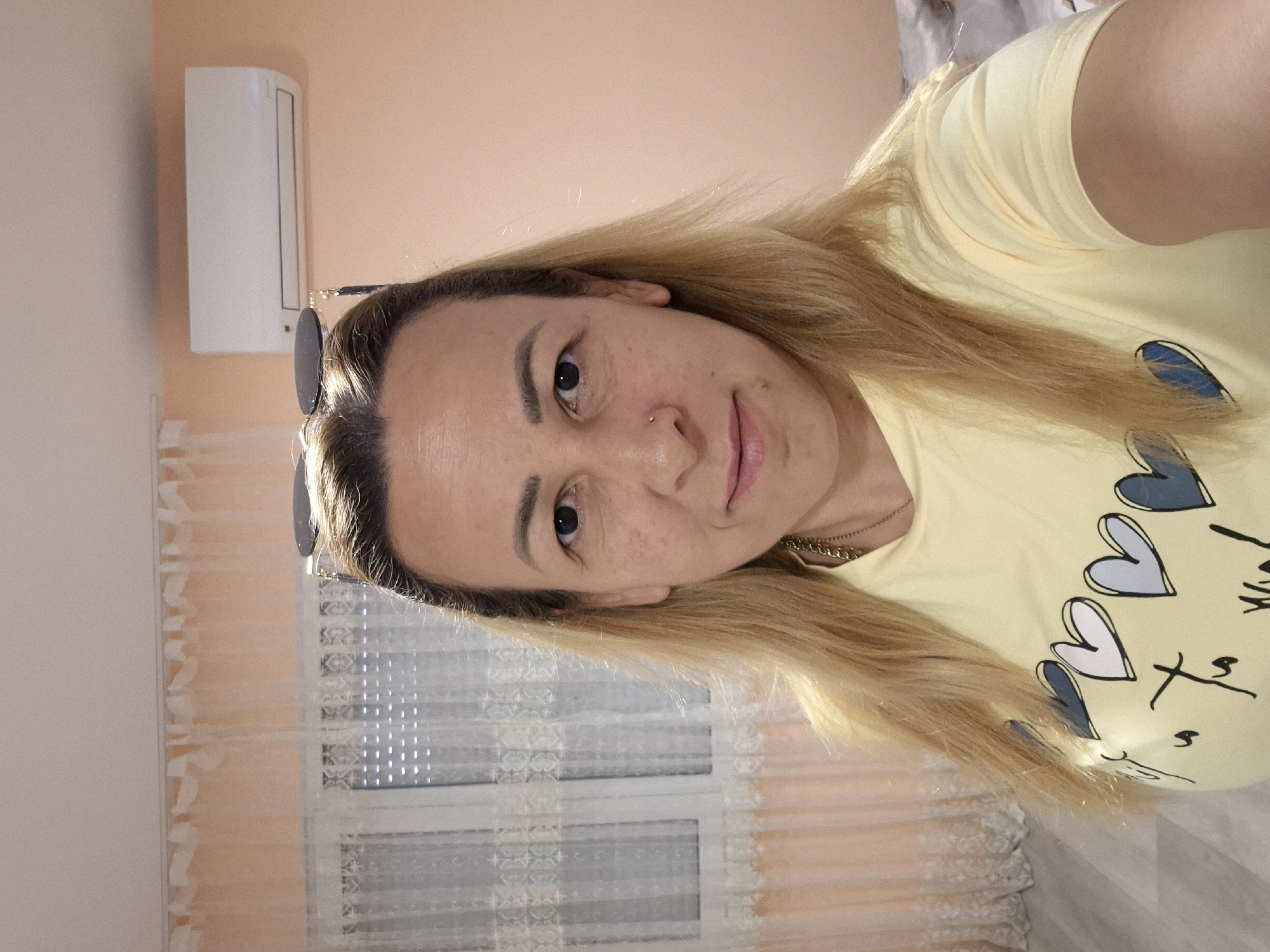 A medium shot shows a blonde woman with fair skin, light brown eyes, and a small nose ring posing for a selfie. She is wearing a light yellow t-shirt with a graphic of hearts and cursive writing. Sunglasses are perched on her head. The background includes a pale pink wall with a white air conditioning unit mounted near the ceiling. To the left, a window with sheer white curtains and blinds is visible. The floor appears to be light-colored wood or laminate. The lighting suggests an indoor, likely residential, setting.