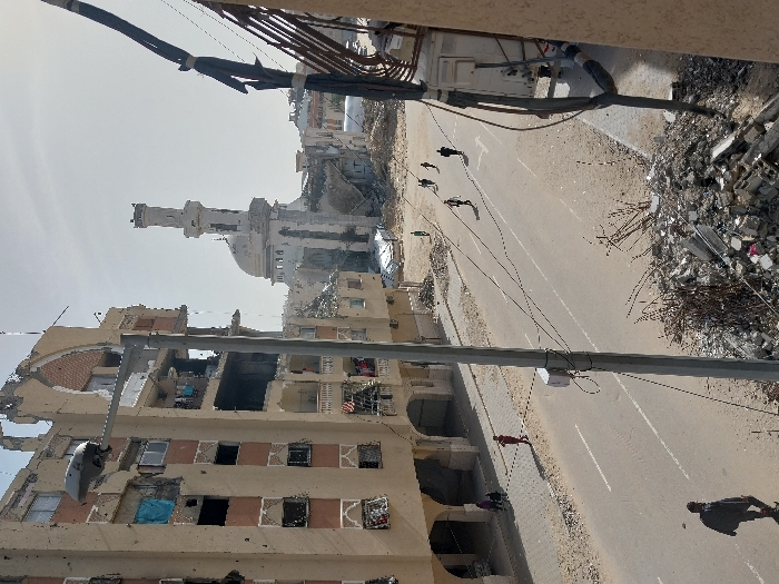 This image captures a scene of extensive urban destruction in the city of Khan Yunis, Palestinian Territory. The view looks down a paved road with faded white lane markings, flanked by rubble and damaged structures.
On the left, a multi-story apartment building is severely damaged, with large sections of its facade missing, revealing exposed interiors, shattered window frames, and visible structural rebar across multiple floors. An arched architectural detail is visible on the upper part of the building. A street light pole stands in front of this building. On the sidewalk below, a group of approximately three individuals walks. Further down the street, another lone individual walks on the left side of the road.
In the mid-ground, a paved street runs diagonally, with several individuals walking across its right lane, moving away from the viewer. Beyond the street, vast areas are reduced to piles of concrete rubble and debris. Dominating the background is a mosque with a distinctive ribbed dome and a tall, slender minaret, both showing signs of structural damage. The sky above is bright but hazy.
In the immediate foreground on the right, a large pile of construction debris, including concrete blocks and twisted rebar, is visible. A thick bundle of black electrical cables hangs vertically from the right edge of the frame.