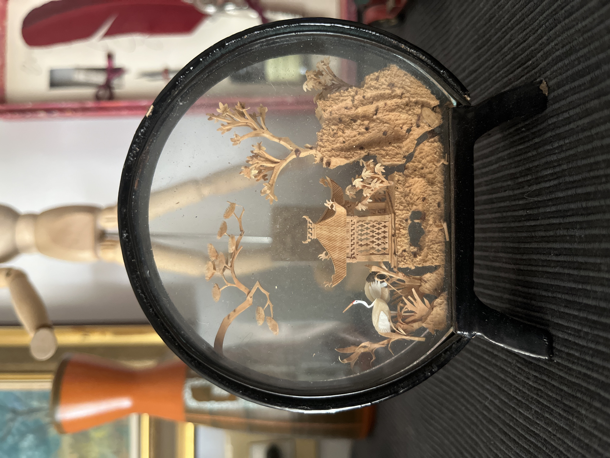This is a close-up, eye-level shot of a circular, black-framed diorama featuring a miniature scene of a pagoda, trees, and a crane. The diorama is supported by two black legs. The background is out of focus and appears to be an indoor setting with other decorative objects, including what looks like a wooden mannequin and a red feather. The lighting is natural, coming from the front, illuminating the diorama and casting soft shadows.