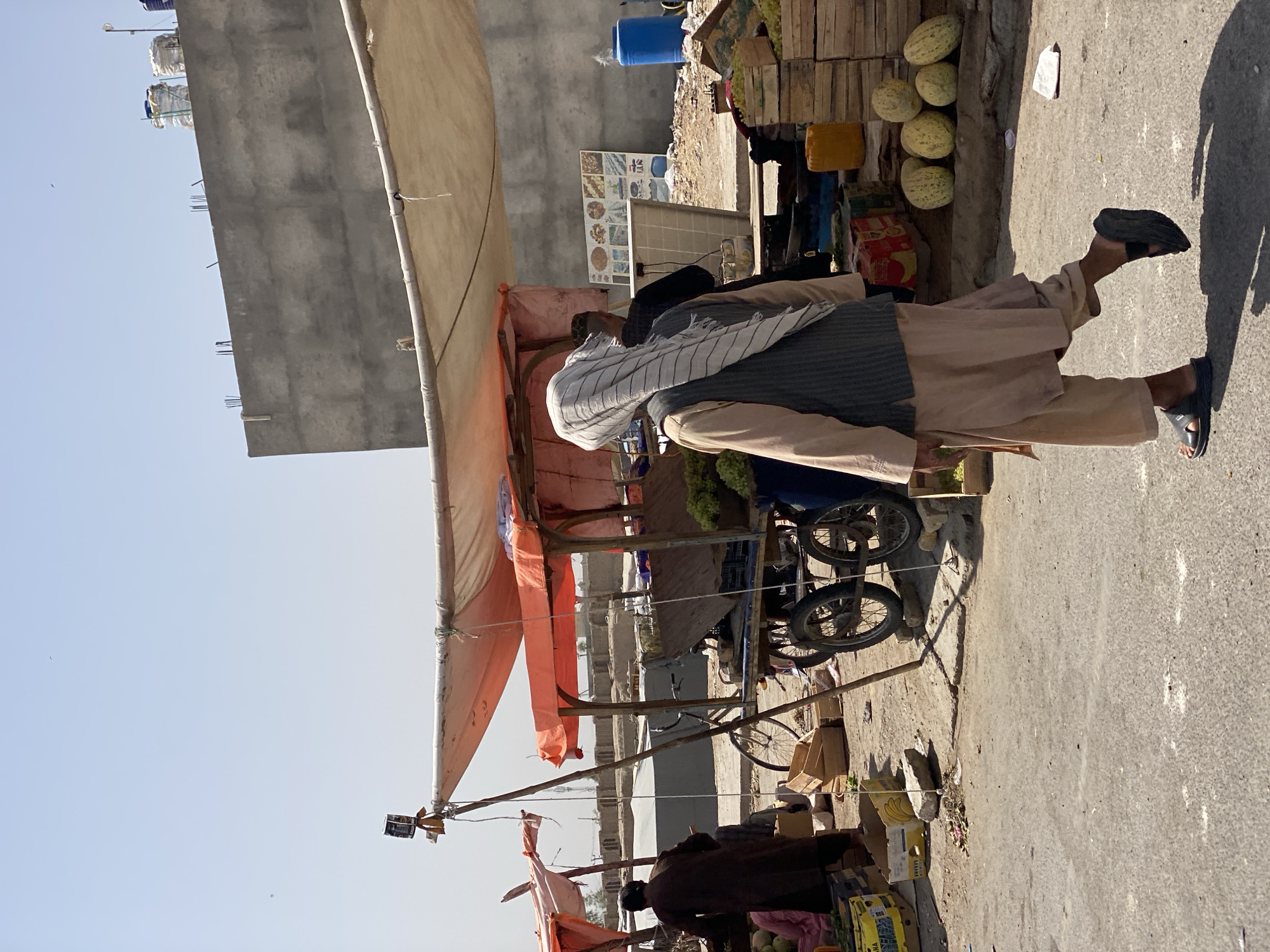 The image captures a street scene in the city of Kandahar, Afghanistan, under a clear, bright sky. In the foreground, a person wearing traditional Afghan clothing, including a light brown shalwar kameez and a grey vest, walks away from the camera. They have a white and grey striped turban wrapped around their head and are wearing dark sandals. To the left, another person in a dark brown robe stands near a cart.
The scene is dominated by market stalls with makeshift canopies, one orange and one a lighter, weathered fabric. The stalls are laden with produce, including what appears to be melons displayed in wooden crates and bunches of green grapes. A bicycle is parked behind one of the carts, and various wooden crates and boxes are scattered around the ground. In the background, a large, unfinished concrete building with rebar protruding from its top looms over the market. Two large blue water tanks and a yellow container are visible near the building. The ground is a dusty, paved surface. The overall impression is of a bustling, open-air market in a developing urban environment.