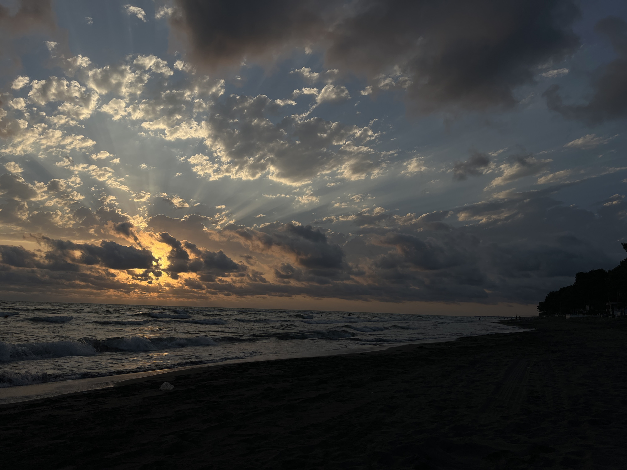 The image depicts a dark, sandy beach with waves crashing onto the shore. The sky is filled with dramatic clouds, with the sun setting or rising behind them, casting rays of light through the clouds. The light from the sun is orange and yellow, illuminating some of the clouds and reflecting on the water. The water is dark and appears to have some choppiness. In the distance, along the coastline, there are trees and some indistinct structures, possibly buildings or beach amenities. There are a few very small figures visible along the waterline and in the water, but their features are not discernible. The overall scene conveys a moody and atmospheric sunset or sunrise over the Black Sea coast in the area of Supsa, Georgia.