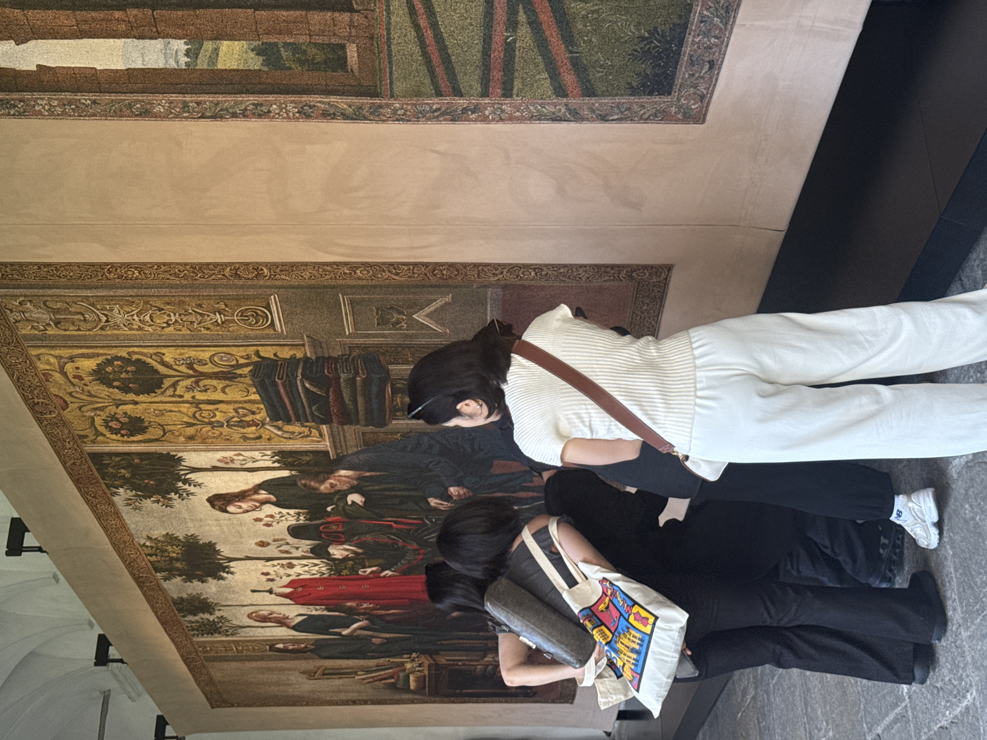 This image, presented rotated 90 degrees counter-clockwise, depicts an indoor scene, likely within a museum or a historic building in Milan, Italy. The setting features a grand wall adorned with a large, elaborate artwork, appearing to be a historical tapestry or fresco. This artwork illustrates a narrative scene with multiple figures dressed in traditional attire, positioned among trees and subtle architectural elements in the background. The piece is framed by intricate decorative borders, which also extend as horizontal patterns along the upper sections of the walls.
Two women are visible, seen from behind or a high angle, observing the artwork. The woman on the right (in the corrected orientation) is dressed in a white, ribbed long-sleeved top, light-colored (cream or white) wide-leg pants, and white sneakers with a discernible blue "B" logo on the side. She carries a brown leather shoulder bag. The second woman, on the left, wears dark clothing and holds a light-colored tote bag featuring a vibrant, abstract graphic design. Both individuals appear focused and absorbed in viewing the art.
The high ceilings and detailed wall decorations suggest a culturally significant venue. The floor is dark, possibly stone or concrete. The lighting is even and bright, typical of an indoor daytime setting. The scene captures the quiet engagement of visitors appreciating historical art in a richly decorated environment. Beyond the shoe logo and the unreadable graphic on the tote bag, no other text is clearly visible.