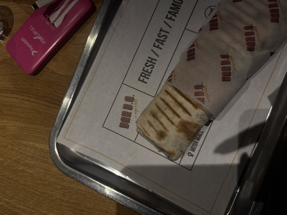 A Bar B.O. angus kebab wrap, marked with grill lines and partially unwrapped, sits on a branded paper. The paper, placed on a metal tray, has the Bar B.O. logo, the phrase "FRESH / FAST / FAMO," and "MEGA MALL." A pink "fastshift POWERFULL" device is positioned on the wooden table alongside the tray. The scene is shot from an overhead perspective.