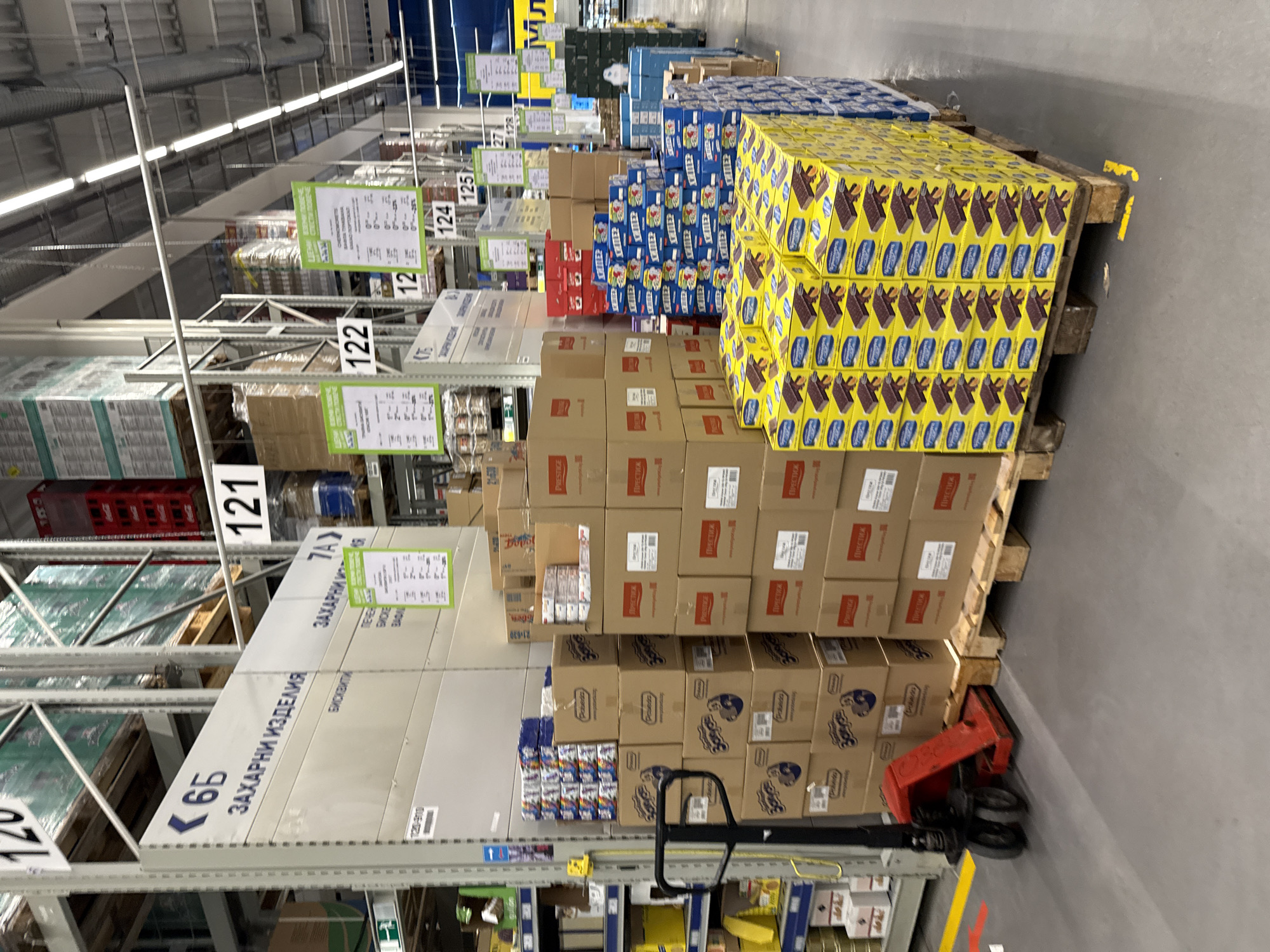 The image depicts the interior of a large retail store, specifically the "Метро" (Metro) store located in Stara Zagora, Bulgaria. Aisles are lined with high shelving units stocked with bulk goods, predominantly stacked boxes of various food products, including boxes of chocolate bars, cookies, and canned goods. Price signs and aisle numbers (6Б, 121, 7А, 122, 12, 124, 125) are visible along the shelving. A red hand pallet jack is stationed near the base of a stack of boxes on a pallet. The overall environment suggests a wholesale or warehouse-style retail setting.