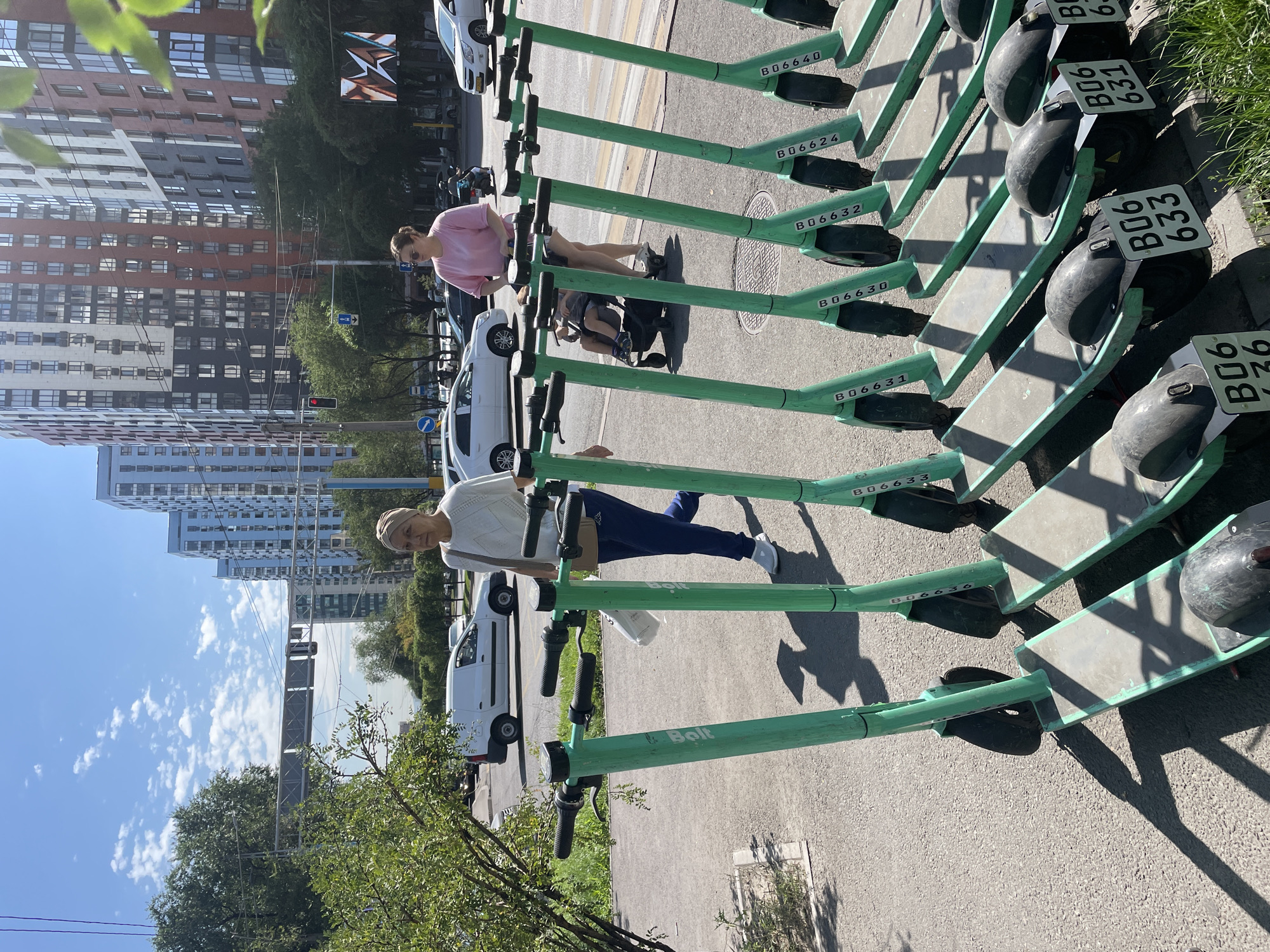 In Almaty, Kazakhstan, on a bright, sunny day, a row of green electric scooters is parked along a sidewalk adjacent to a street. The scooters have the "Bolt" logo on their handlebars. In the background, modern, multi-story apartment buildings with colorful facades and numerous windows dominate the skyline. A few cars, including a white van, are visible on the street, and traffic lights are present. Two women are walking on the sidewalk. One woman, wearing a white top and dark blue pants, is walking away from the camera, carrying a white bag. The second woman, wearing a pink shirt and shorts, is walking towards the camera, pushing a stroller with a child in it. The scene captures a typical urban environment with elements of modern architecture and common modes of transportation.