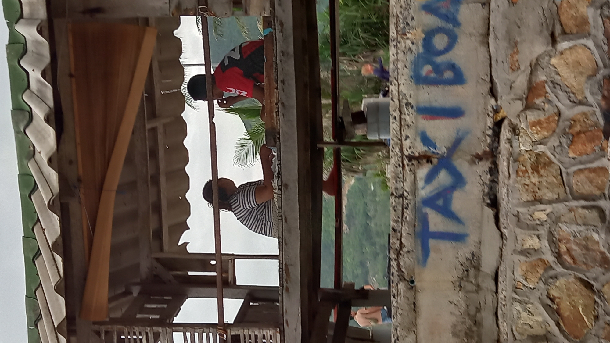 The image displays a multi-layered scene in ตำบลเกาะพะงัน, Thailand, under an overcast sky.
In the foreground, a concrete wall with visible cracks and wear is present, topped by irregular stone masonry. Blue spray paint on the concrete spells "TAXI BOAT," with the final "T" partially obscured.
Above this wall, an elevated, open-sided wooden structure houses two individuals. The structure features a corrugated roof, part green and part grey, supported by wooden beams.
- On the left, a female with dark hair, pulled back, is seated in profile. She wears a horizontally striped t-shirt and rests her forearms on a wooden surface in front of her. Her right foot, wearing an orange or red sandal, is visible dangling below the platform.
- On the right, a male is seated in profile, facing the female. He wears a red and black short-sleeved top with a white design (possibly "2N") on the right shoulder/sleeve. He holds a small, light-colored object to his mouth.
- The two individuals are situated across a wooden surface or railing.
Through the open sides of the structure, lush green foliage, including palm fronds, is visible. Beyond this, distant, tree-covered hills rise against the cloudy sky.
Below the main platform of the elevated structure, on a lower level or ground, a partially visible male figure wearing a light-colored shirt and blue shorts stands on the far left. Further to the right, another small figure in blue/purple is barely discernible in the middle ground, behind the foreground wall.