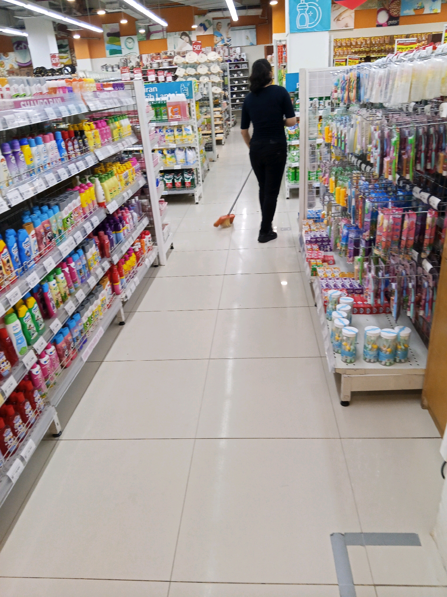 The image captures an indoor scene, likely a retail environment such as a supermarket or department store, in Lubuklinggau, Indonesia. A woman is seen from behind, dressed in a black top and black pants, pushing a broom across the tiled floor. She is positioned in the center-right of the frame, walking away from the camera. To her left, shelves are stocked with various consumer goods, including bottles of liquids, likely cleaning supplies or personal care items. To her right, more shelves display rows of packaged products, possibly toiletries or household items. The aisle is well-lit by overhead fluorescent lighting. The floor is composed of large, light-colored square tiles with grout lines visible. A grey tape marking is present on the floor in the lower right corner of the image. The overall impression is of a clean, organized commercial space.