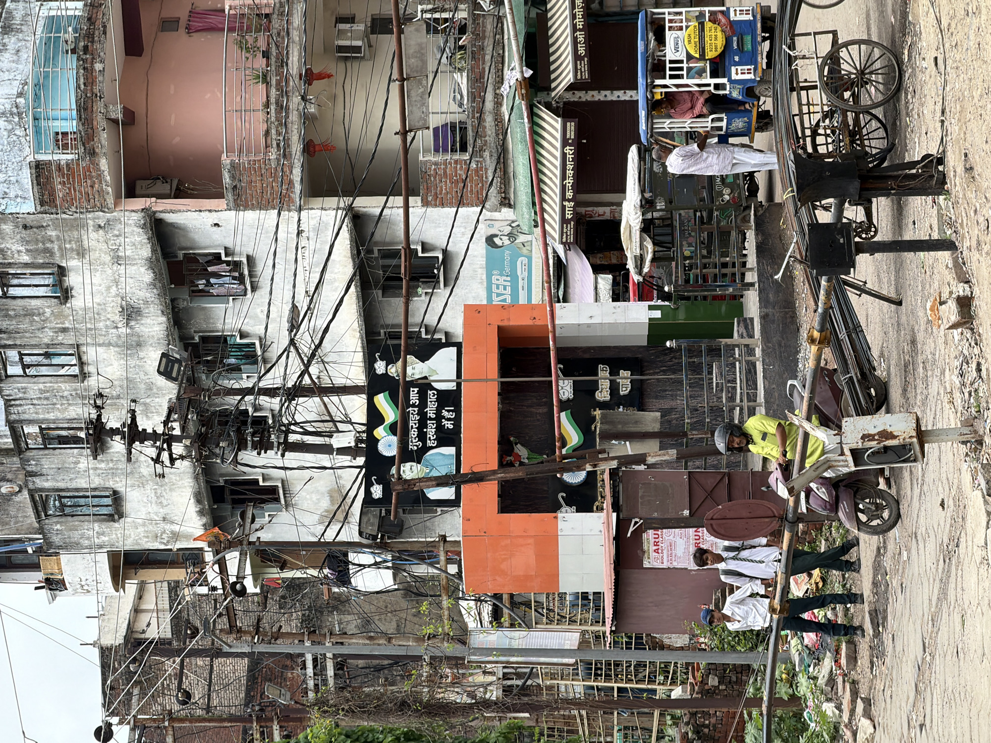 The image depicts a street scene in Kanpur, India. A tall, multi-story building with weathered, concrete walls dominates the background. The building has numerous windows, some of which are open, revealing laundry hanging inside. A complex network of electrical wires and poles crisscrosses the foreground and mid-ground, indicative of densely populated urban areas.
In the mid-ground, an orange and white structure features a large black banner with images of two men and Indian flags. Text in Hindi is also visible on the banner. To the left of this structure, a younger man in a light-colored shirt and dark pants stands next to another young man in a similar outfit, wearing a blue cap. They appear to be standing near a gate or railing.
In the foreground, a man in a bright yellow shirt and a helmet is seated on a scooter. The scooter is parked on a dusty street. Beside the scooter, a wooden cart is loaded with metal rods. A blue and white auto-rickshaw is visible to the right, with two men inside. The environment appears somewhat cluttered, with debris and rubble on the side of the road. The sky is overcast, suggesting a cloudy day.