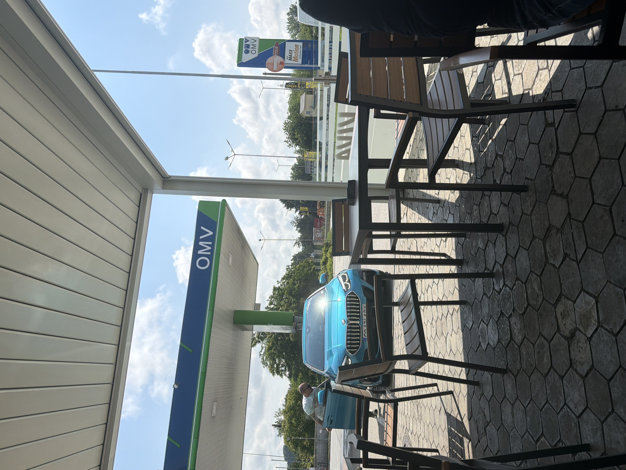 An outdoor scene depicts a gas station with an adjacent seating area under a clear, sunny sky, suggesting daytime. The main subject is an OMV gas station, identifiable by its prominent green and blue logo on a tall sign and the station canopy.
In the midground, a striking bright blue car, possibly a BMW, is parked at a fuel pump under the OMV canopy. A man, appearing bald and wearing a light-colored shirt, is captured in the act of opening or closing the driver's side door of the blue vehicle. The car's license plate, "CA 1744 DV," is visible.
To the right of the frame, in the foreground, is an outdoor seating area furnished with several dark metal tables and chairs featuring wooden slats. Sunlight casts distinct shadows across the patterned ground, which is paved with hexagonal blocks.
In the background, beyond the gas station, there are green trees and a bright blue sky dotted with scattered white clouds. A tall OMV sign also displays "VIVA" and "MAXXMotion" logos, alongside what appear to be fuel prices: "1.65" and "1.74". A red circular "no entry" or "forbidden" sign is also visible on this pole. The overall setting suggests a typical roadside stop or service area.