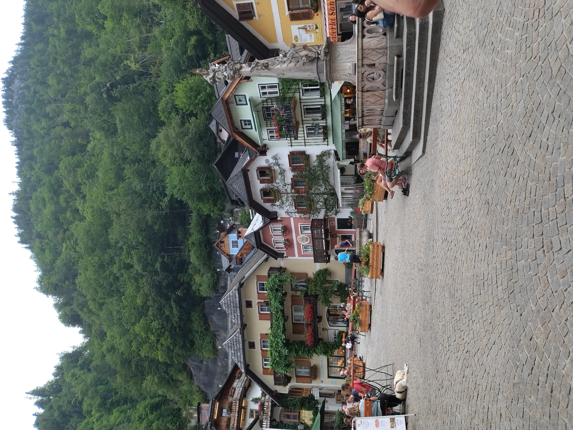 This is a vertical, eye-level, medium-shot photograph of the Marktplatz in Hallstatt, Austria. The scene is dominated by the architecture of the town, with colorful buildings featuring wooden balconies, shutters, and pitched roofs, set against a backdrop of a steep, densely forested mountainside.
In the foreground, a cobblestone plaza is visible, with several outdoor seating areas for cafes and restaurants. A white dog is lying down on the cobblestones near a table. A few people are seated at tables, and others are walking through the plaza.
A prominent feature in the middle ground is a stone fountain with a statue on top. The fountain is surrounded by steps and is a focal point of the plaza.
The buildings surrounding the plaza are a mix of styles and colors, including yellow, cream, pink, and green. Many of the buildings have window boxes filled with flowers, adding pops of color to the scene. Signs for businesses are visible on some of the buildings, such as "Salzwiener" and "Hotel Bar".
The overall atmosphere appears to be one of a vibrant European town square, likely during daytime, with a mix of locals and tourists enjoying the surroundings. The lush green mountainside provides a dramatic and picturesque backdrop to the historic town.