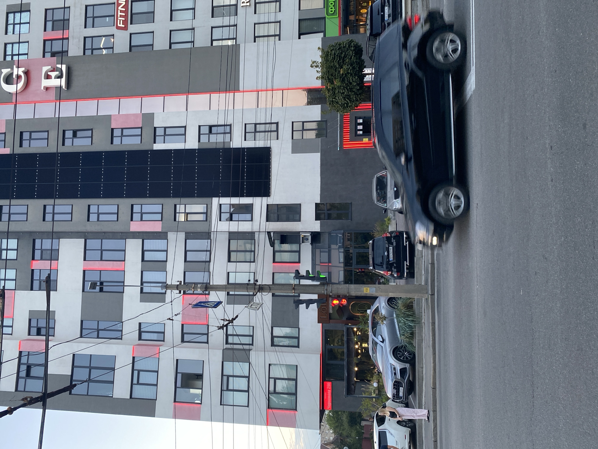 The image depicts a street scene in Vinnytsia, Ukraine, with a modern high-rise building as the prominent feature. The building is characterized by its grey and white facade, interspersed with red and pink rectangular lighting elements. Large windows with dark frames are visible across the building's exterior. The top of the building displays large white letters "G E". A sign with the word "FITNI" is visible on the right side of the building.
In the foreground, a black SUV is in motion, blurred due to its speed, crossing the street. To the left of the moving vehicle, several parked cars are visible, including a silver Audi SUV and a white electric car. A woman in a black top and pink pants stands on the sidewalk near the parked cars.
A traffic light is visible on a utility pole, showing a green light. A pedestrian crossing sign is also present on the same pole. The street is paved with asphalt. Businesses with awnings and illuminated signs, including "LÖLİ" and "Чорновола 298", are located at the base of the building. Trees and greenery are present near the building's entrance and along the sidewalk. Overhead wires cross the scene, suggesting an urban environment with established infrastructure. The sky appears overcast.