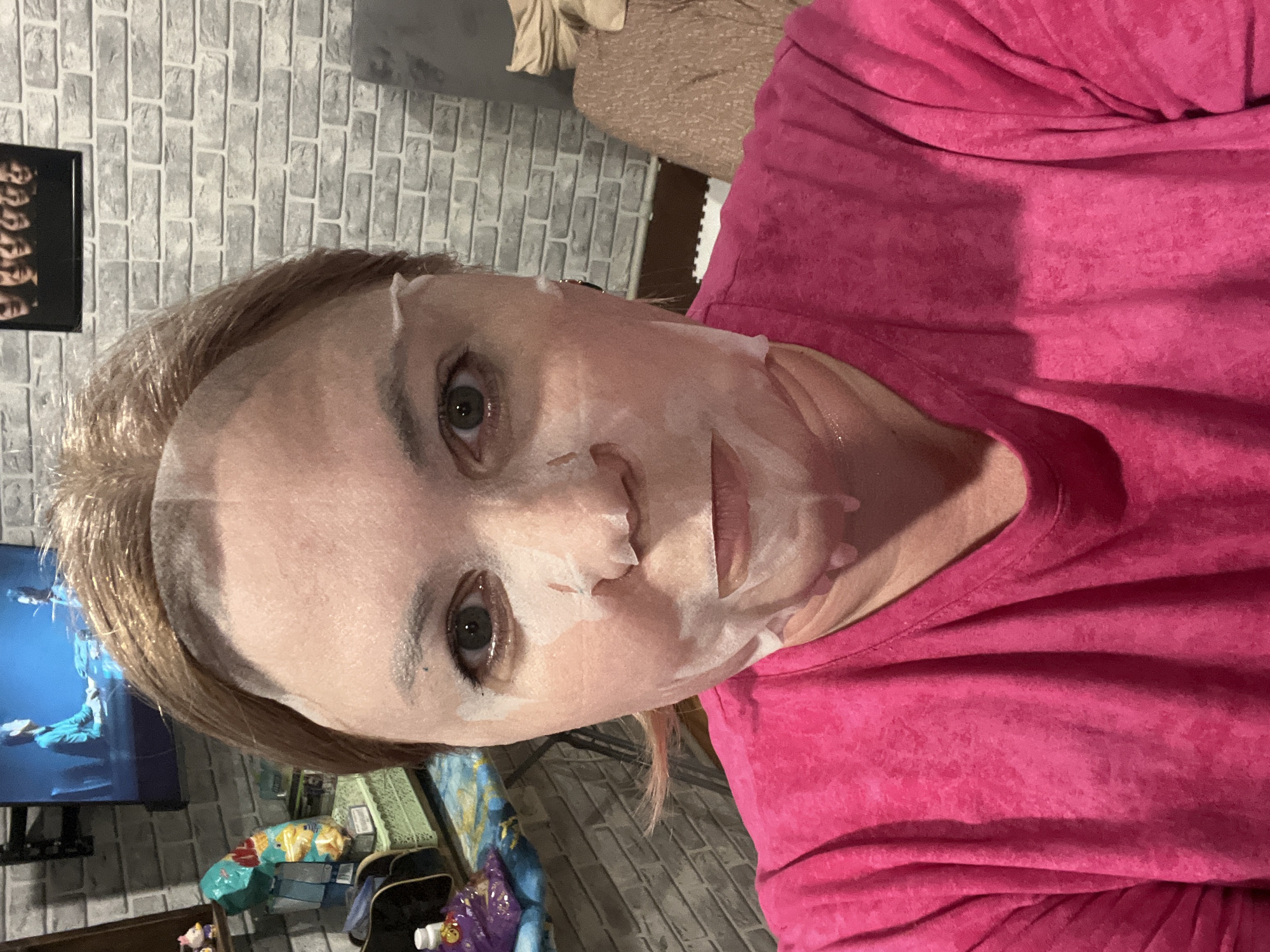 A woman is facing the camera, wearing a pink shirt and a white sheet mask on her face. The background includes a brick wall, a television screen showing an image of people, a shelf with various items, and a portion of what appears to be a couch or bed. The location is not identifiable as Vesyoloye, Russia.