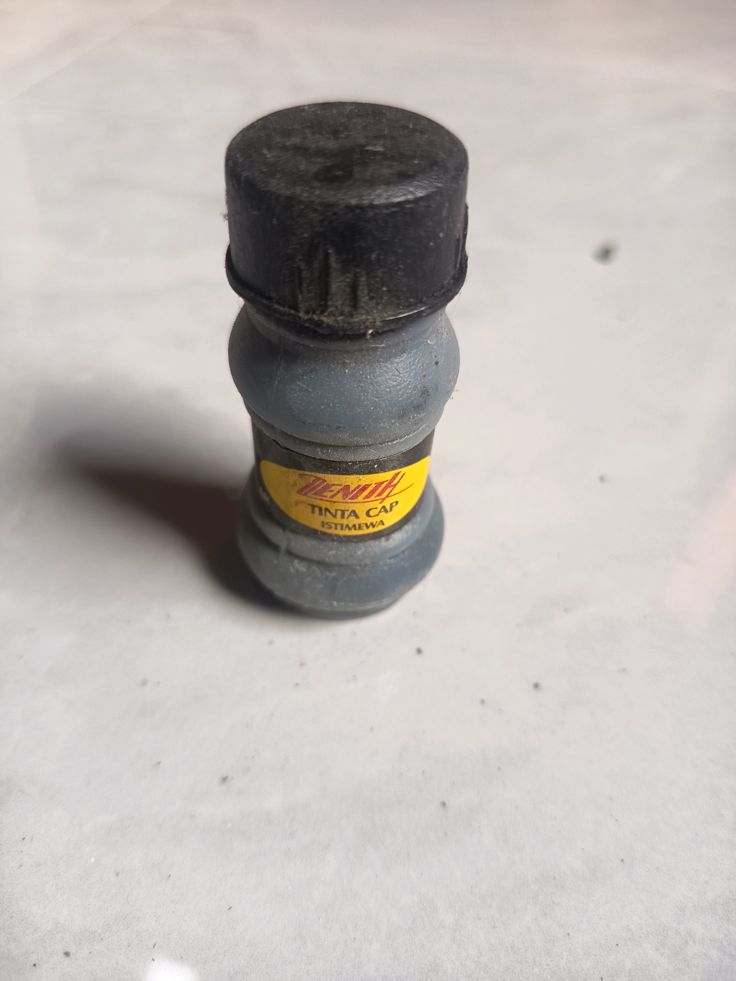 A small, gray bottle with a black cap stands on a tiled surface. The bottle has a yellow label that reads "ZENITH TINTA CAP ISTIMEWA". The tiled surface appears to be outdoors and is slightly dusty. The lighting suggests it is daytime. The location of this scene is within the city of Sidorejo, Indonesia.