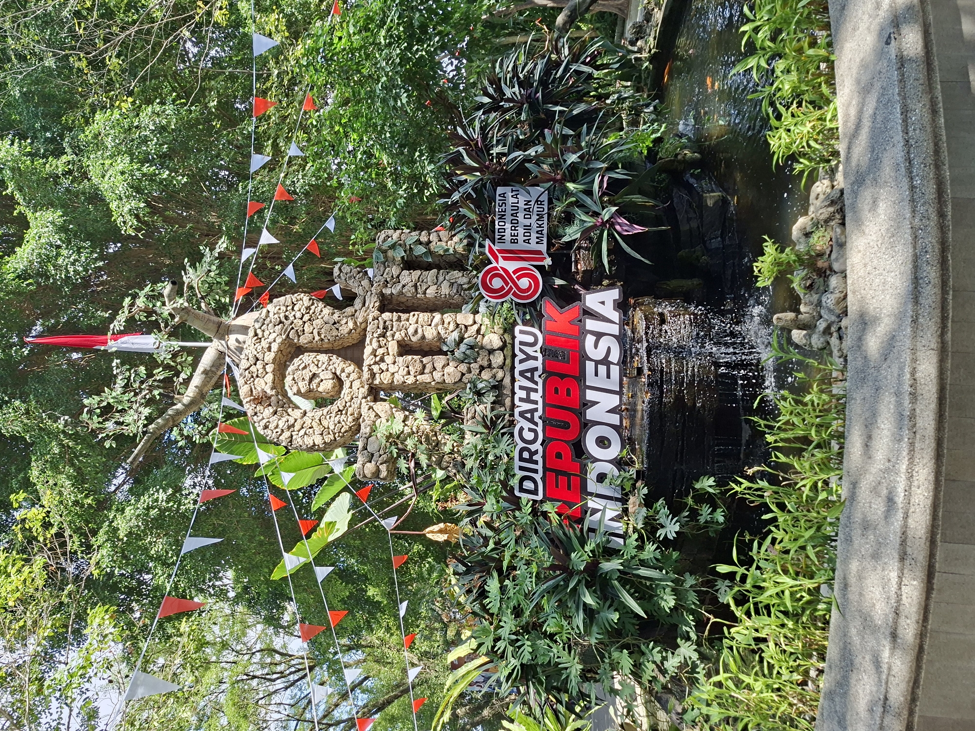 The image depicts a decorative fountain and monument celebrating Indonesia's Independence Day, likely in the city of Yogyakarta, Indonesia, given the context of national holidays and the prevalent Indonesian language on signage. The central element is a stone sculpture resembling the number "81", adorned with a flag of Indonesia (red and white) at its apex. Below the sculpture, a large sign reads "DIRGAHAYU REPUBLIK INDONESIA" in bold red and white lettering. To the right of this sign, another smaller sign displays text in Indonesian: "INDONESIA BERDAULAT ADIL DAN MAKMUR" which translates to "Sovereign, Just, and Prosperous Indonesia". The fountain features cascading water and lush green foliage, including various tropical plants and some bamboo-like stalks in the foreground. A small pond with visible orange koi fish is present to the right of the fountain. Festive red and white triangular pennant banners are strung across the scene, adding to the celebratory atmosphere. The background is dominated by the dense green canopy of large trees, suggesting an outdoor location possibly within a park or garden. No people are visible in the frame.
