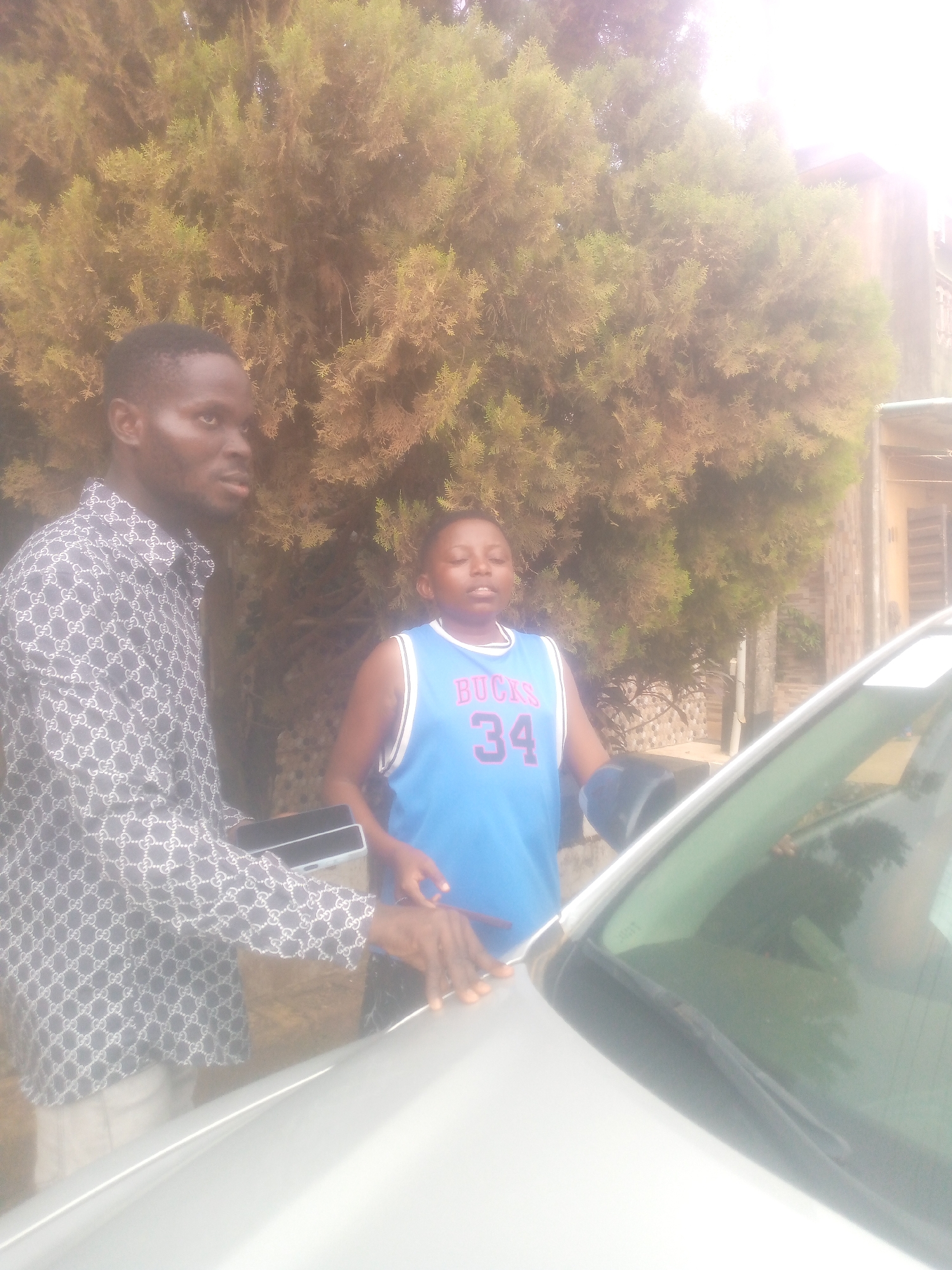 A medium shot reveals two males standing near a silver car parked in front of a residence in an unknown city. The older male on the left is wearing a patterned shirt and holding a black phone. He is standing with his hand on the vehicle's hood. The younger male on the right is wearing a blue sports jersey with the word "BUCKS" and the number "34" visible on the front. A large tree is behind them. The building behind the tree, along with the car, are bathed in sunlight.