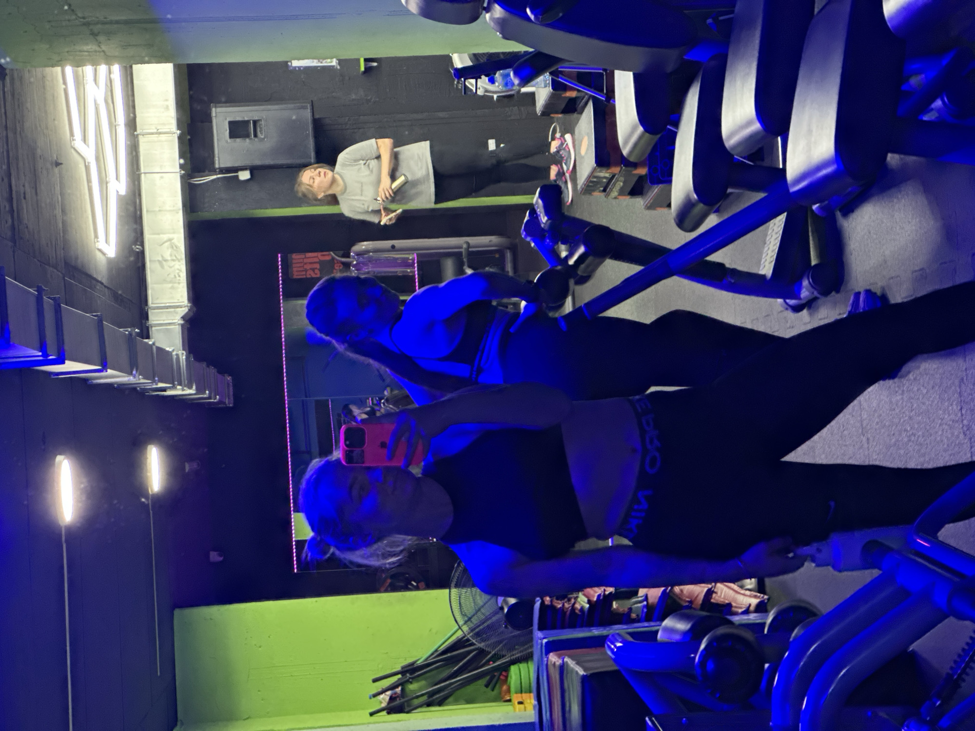 The image depicts a woman taking a selfie in what appears to be a gym, possibly in Budva, Montenegro. The gym has exercise machines with blue accents. Two other people can be seen in the background, one using a machine and another standing near a water bottle. The gym has green walls with lights and architectural details.