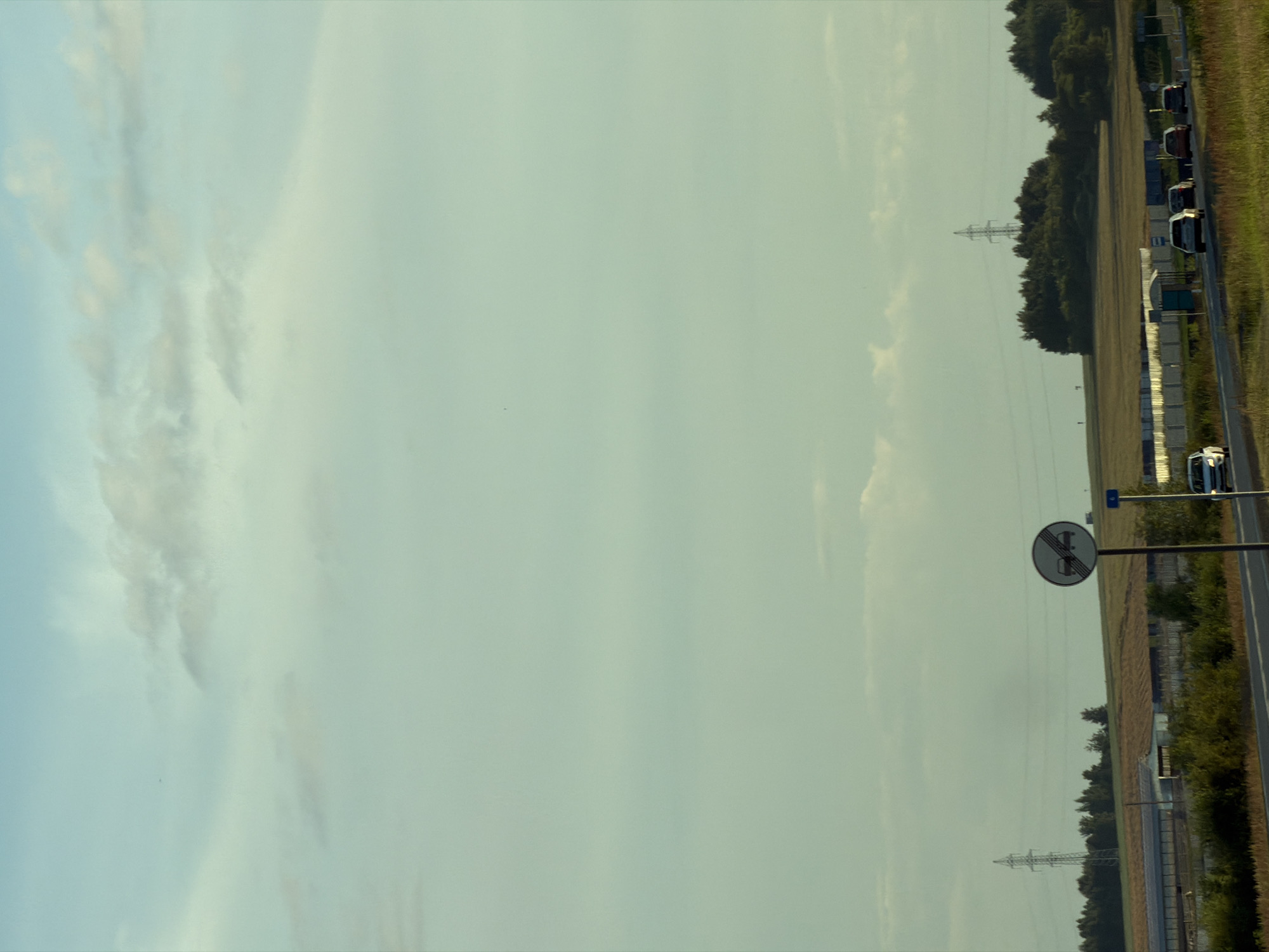 The image shows a rural scene with a road, cars, and a traffic sign. In the foreground, a road curves to the right, with several cars driving on it. A circular traffic sign with a red border and two red cars inside, indicating "no overtaking," is positioned on a pole to the right of the road. In the background, there are fields, trees, and a power line tower. The sky is light blue with some clouds. The overall impression is one of a sunny day in a rural setting.