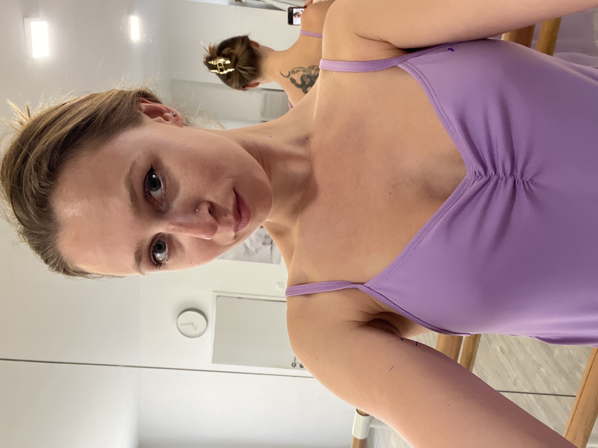 The image captures a woman taking a selfie in a brightly lit indoor setting, likely a dance studio or practice room in Budva, Montenegro. The primary subject is a light-skinned woman with light brown hair pulled back, wearing a purple leotard or swimsuit with thin straps and a ruched bust. She has a subtle nose piercing and distinct eyeliner. Her head is tilted, and she looks directly into the camera with a neutral, calm expression.
The background is dominated by a large mirror, reflecting the woman's back and the room behind her. In the reflection, her hair is visible in a bun secured with a golden claw clip, and a dark tattoo of a stylized creature or figure is prominent on her left shoulder blade. Her arm, holding a smartphone to take the picture, is also visible in the reflection.
The room features white walls, two distinct rectangular light fixtures, and a minimalist white circular wall clock. A wooden ballet barre or the back of a wooden chair is partially visible in the lower right, reinforcing the studio environment. The lighting is even and bright, suggesting an interior setting with ample illumination. No visible text is present in the image.