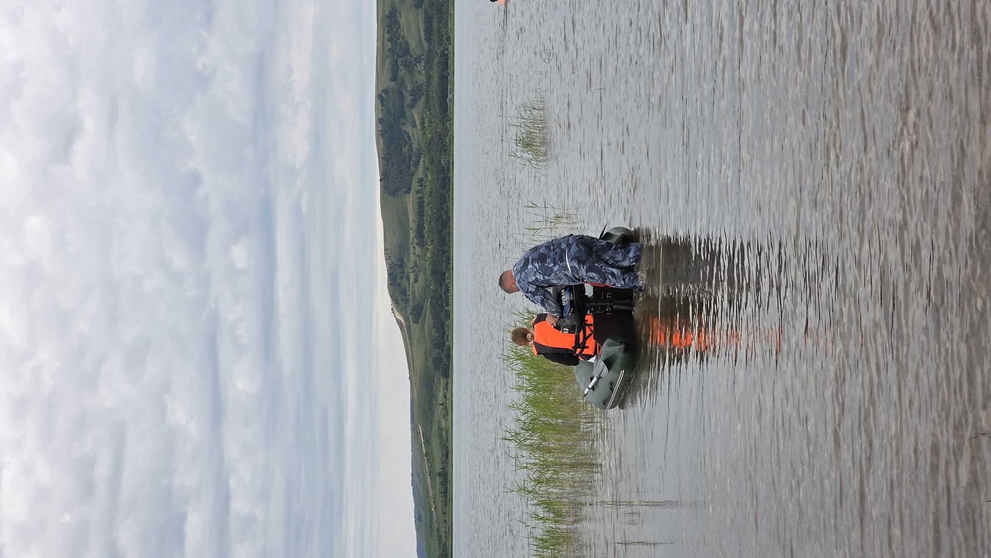 Two individuals are in a dark green inflatable boat on a body of water. One person, wearing an orange life vest over dark clothing, is seated in the boat facing away from the camera. The second person, wearing a camouflage uniform, is standing in the water behind the boat and appears to be adjusting or operating a small outboard motor attached to the stern. The boat is partially submerged, indicating it may be near the shore or in shallow water. Tall green reeds are visible along the left edge of the water, extending towards the boat. The background features a wide expanse of water, with a gently sloping, treed hillside in the distance under a cloudy sky. A road is visible winding its way up the hillside. The overall scene suggests an outdoor activity on a lake or large river, possibly related to recreation or work.