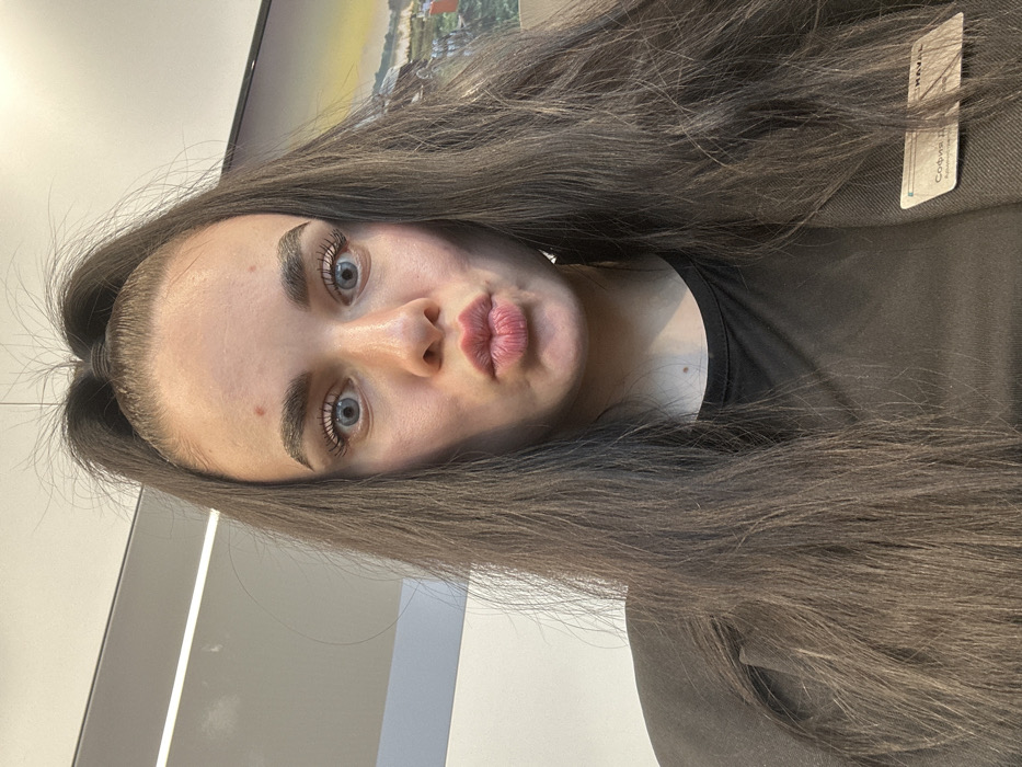 A young adult female is depicted in a close-up, chest-up indoor portrait. She has long, dark brown hair styled in a high ponytail, with some loose strands framing her face. Her eyes are wide open and light-colored, while her lips are tightly pursed in a forward-projecting "duck face" expression. Her eyebrows are dark and prominent. She has visible minor skin blemishes on her forehead and neck. She is wearing a dark short-sleeved top beneath a dark blazer or jacket. An identification badge is clipped to the left side of her chest, displaying the Russian name "София Д." (Sofia D.), the title "Ассистент-рекрутер" (Assistant Recruiter), and a partial "HAVAL" logo. The background consists of a light-colored wall or partition on the left, with a linear ceiling light fixture. To the right, a framed image or digital display shows a landscape scene featuring water, greenery, and structures. The identification badge's text and HAVAL logo indicate the scene is likely within a HAVAL automotive dealership or office in Russia, consistent with HAVAL's operations in the city of Saint Petersburg.