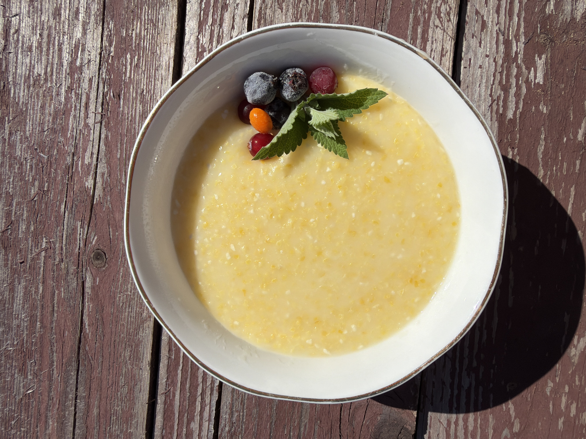 An overhead, medium shot shows a white bowl of yellow corn porridge topped with a sprig of mint and several berries, including frozen blueberries, red currants, and an orange berry, positioned on a weathered, dark red wooden picnic table in Chaikovsky, Russia. The porridge has a creamy texture with visible flecks of cornmeal and some white curds. The berries are clustered on the right side of the bowl, with the mint sprig extending into the porridge. Sunlight casts a strong shadow to the left of the bowl, indicating an outdoor setting. The wooden surface of the table exhibits visible grain and peeling paint, adding to its rustic appearance. No people are visible in the image.