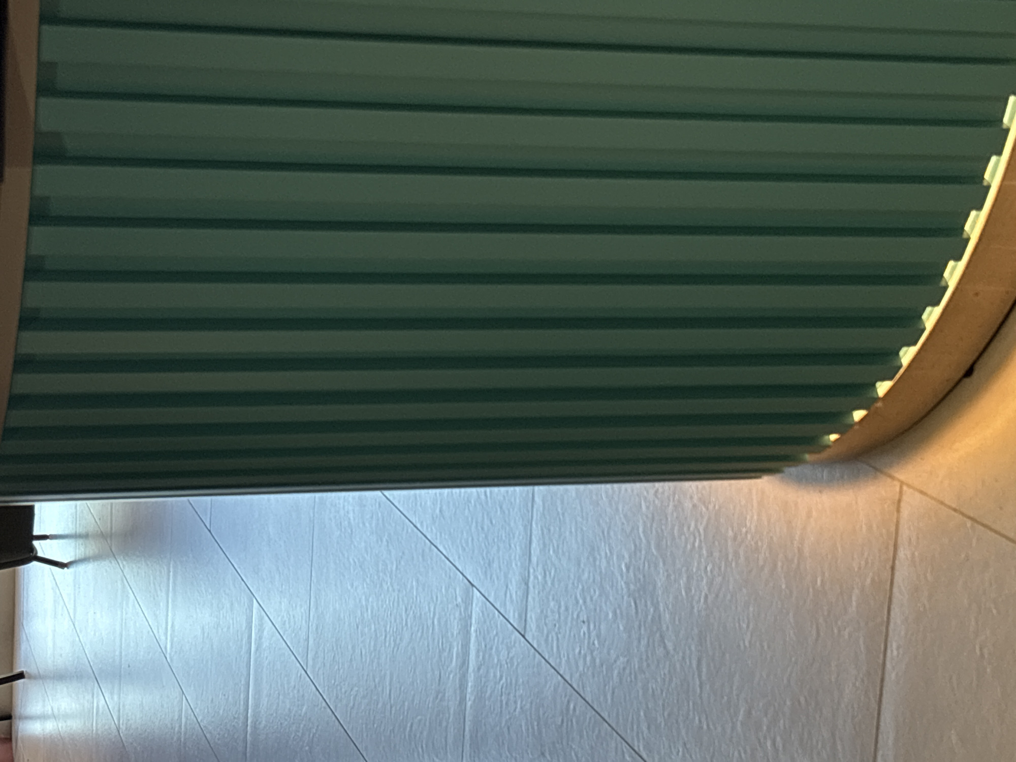 The image shows a close-up view of a teal-colored, ribbed cylindrical structure with a light-colored, possibly wooden base, illuminated by a warm light from below. The structure is positioned on a tiled floor with visible grout lines. The tiles are light beige and laid in a diagonal pattern. In the background, there is a hint of furniture legs and a shadow, suggesting an indoor environment. There are no people visible in the image. The scene appears to be a modern interior, possibly a cafe, bar, or reception area, located in Saint Petersburg, Russia.