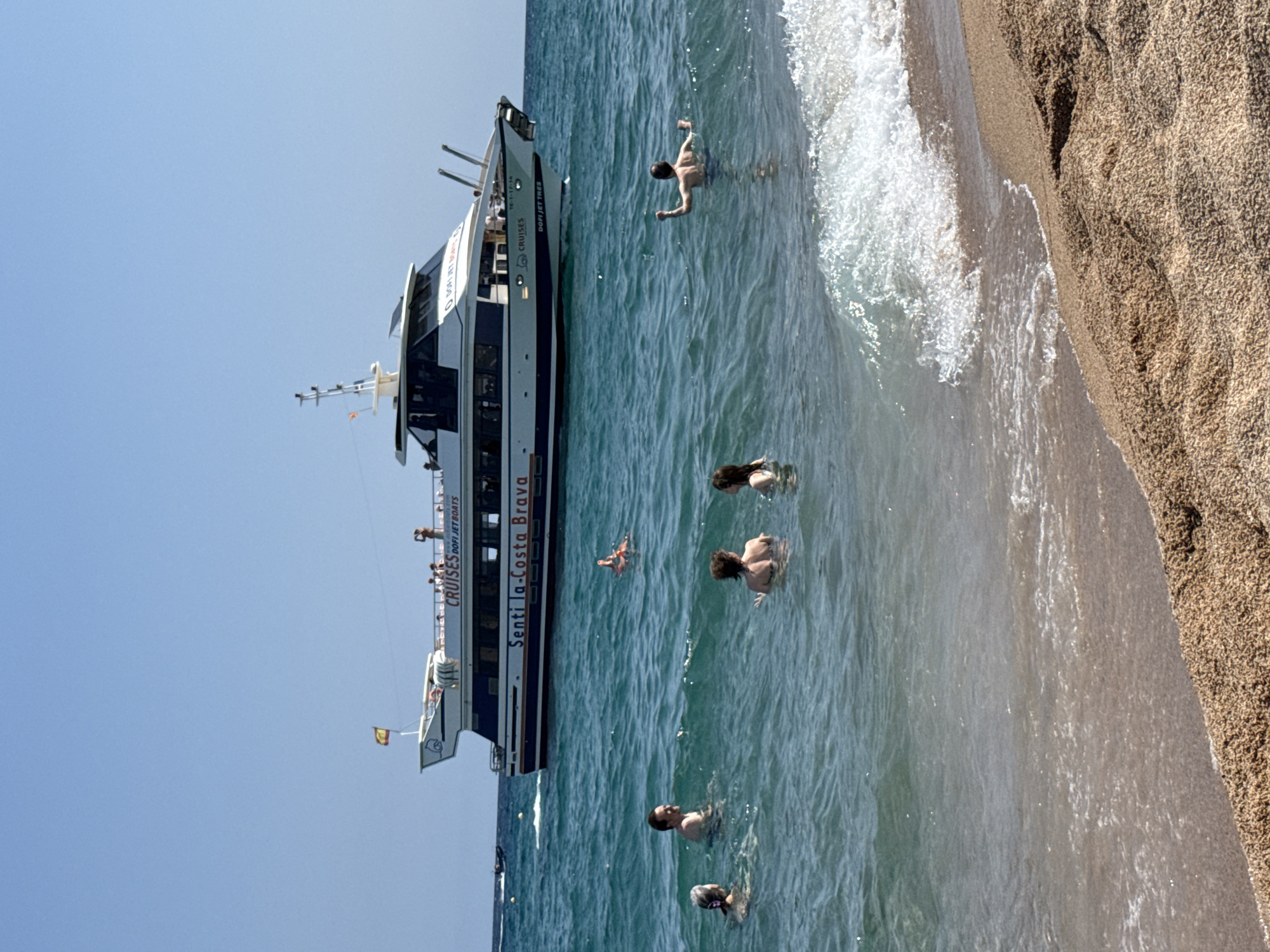 A group of people are swimming in the ocean near a beach with a large boat in the background. The boat, labeled "Senti la-Costa Brava," is a white passenger ferry. A Spanish flag flies from the boat's mast. Several people are visible on the deck of the boat. In the foreground, five people are in the water. One person is floating on their back on a flotation device. The water is clear and blue, with small waves lapping at the sandy shore. The sky is clear and blue. The scene appears to be a sunny day at the beach in Blanes, Spain.