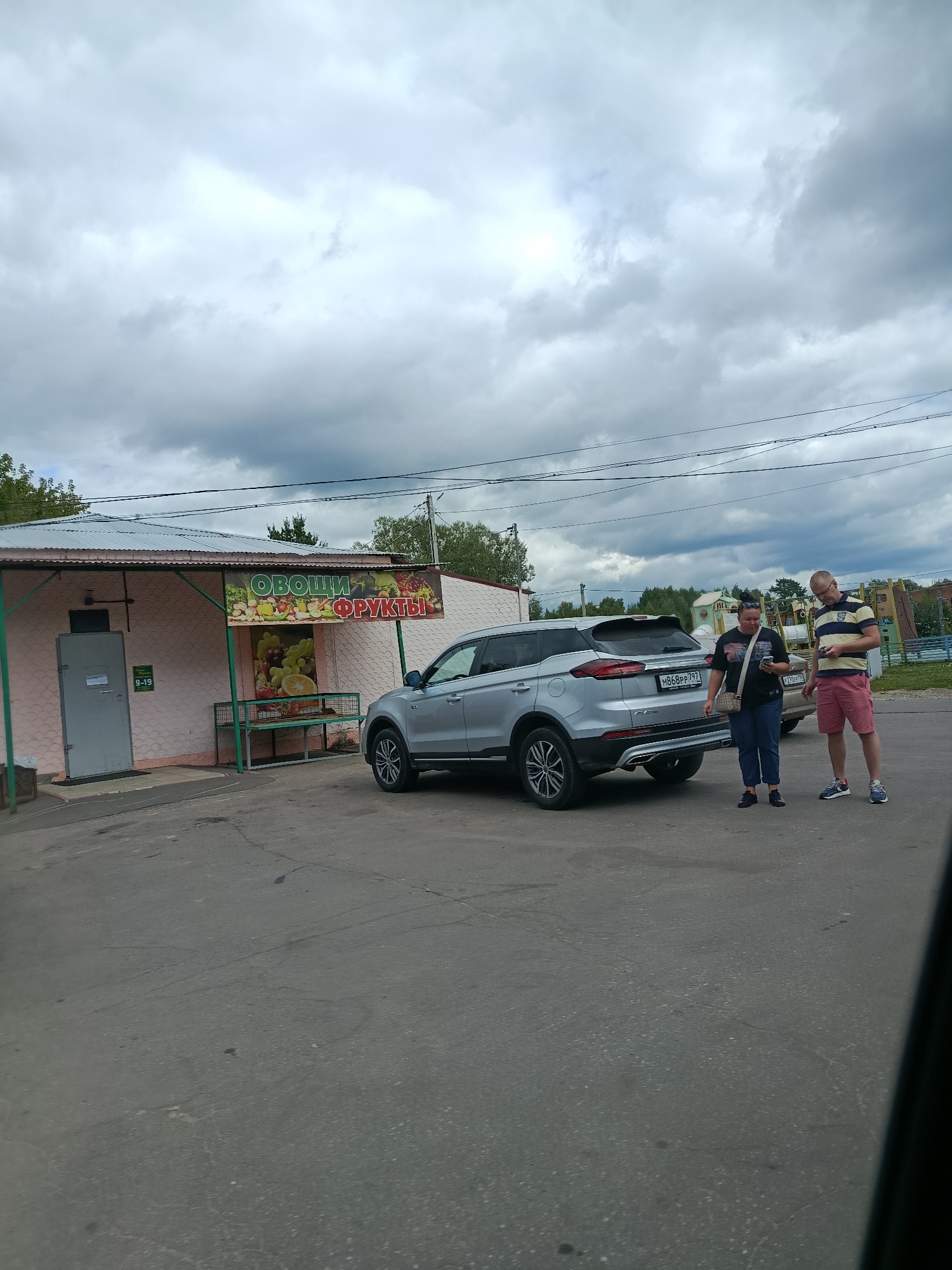A silver SUV is parked in front of a small, light pink building that appears to be a fruit and vegetable stand, indicated by a banner above the entrance that reads "Овощи Фрукты" (Vegetables Fruits). A sign on the door states "9-19." Two people, a woman and a man, are standing near the rear of the SUV. The woman is wearing a black t-shirt and blue jeans, with a beige purse slung over her shoulder. The man is wearing a striped t-shirt and red shorts. The ground is paved with asphalt. In the background, there are trees, a playground structure, and residential buildings. The sky is overcast with heavy clouds. The location is identified as Veriya, Russia.