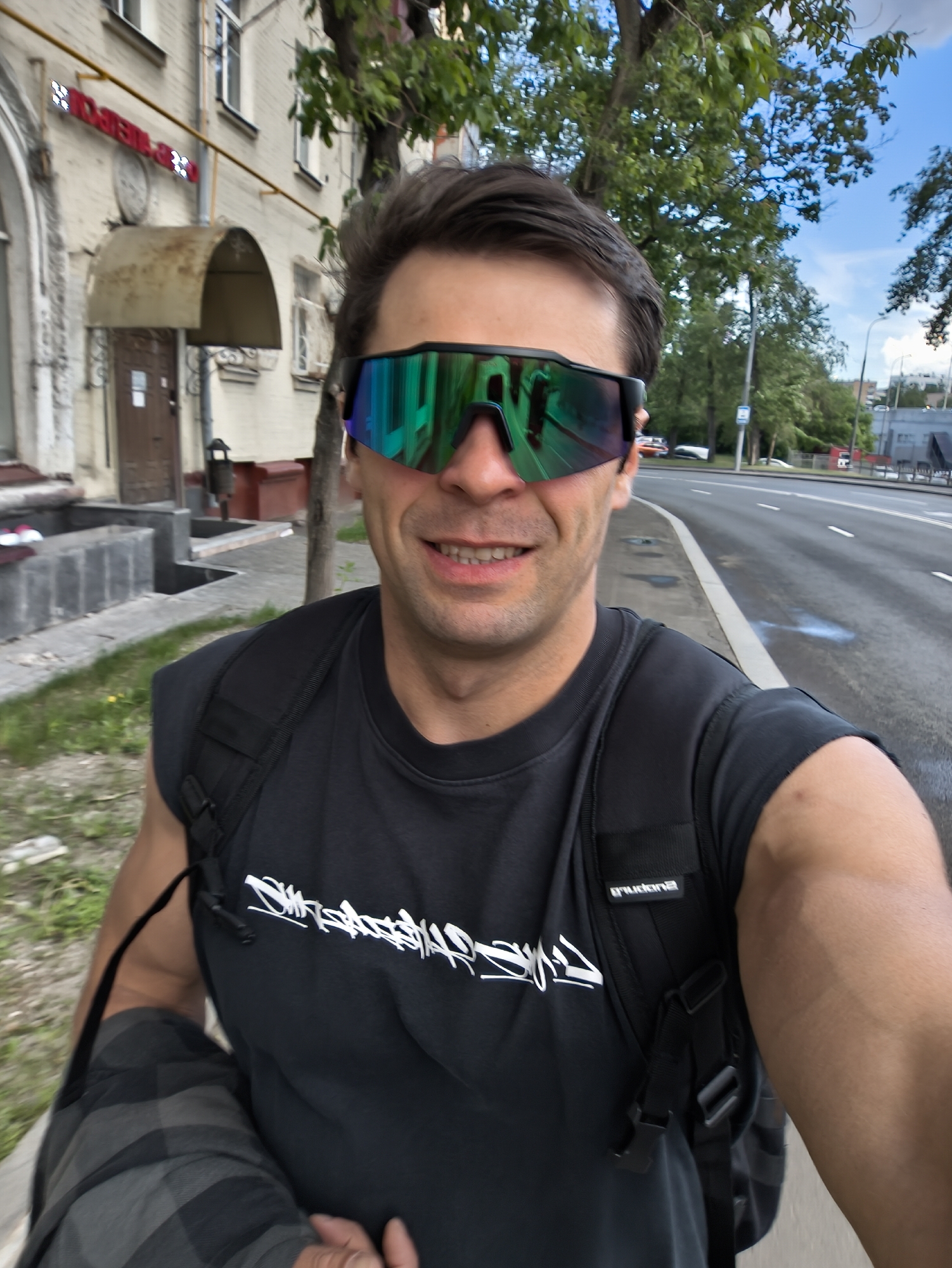A man wearing sunglasses, a dark gray t-shirt, and a black backpack is standing on a sidewalk next to a city street. He is holding a gray checkered jacket in his left hand and is looking at the camera with a slight smile.
In the background, there is a building with a metal canopy and a sign in Russian, trees, and a street with cars. The location appears to be Moscow, Russia.