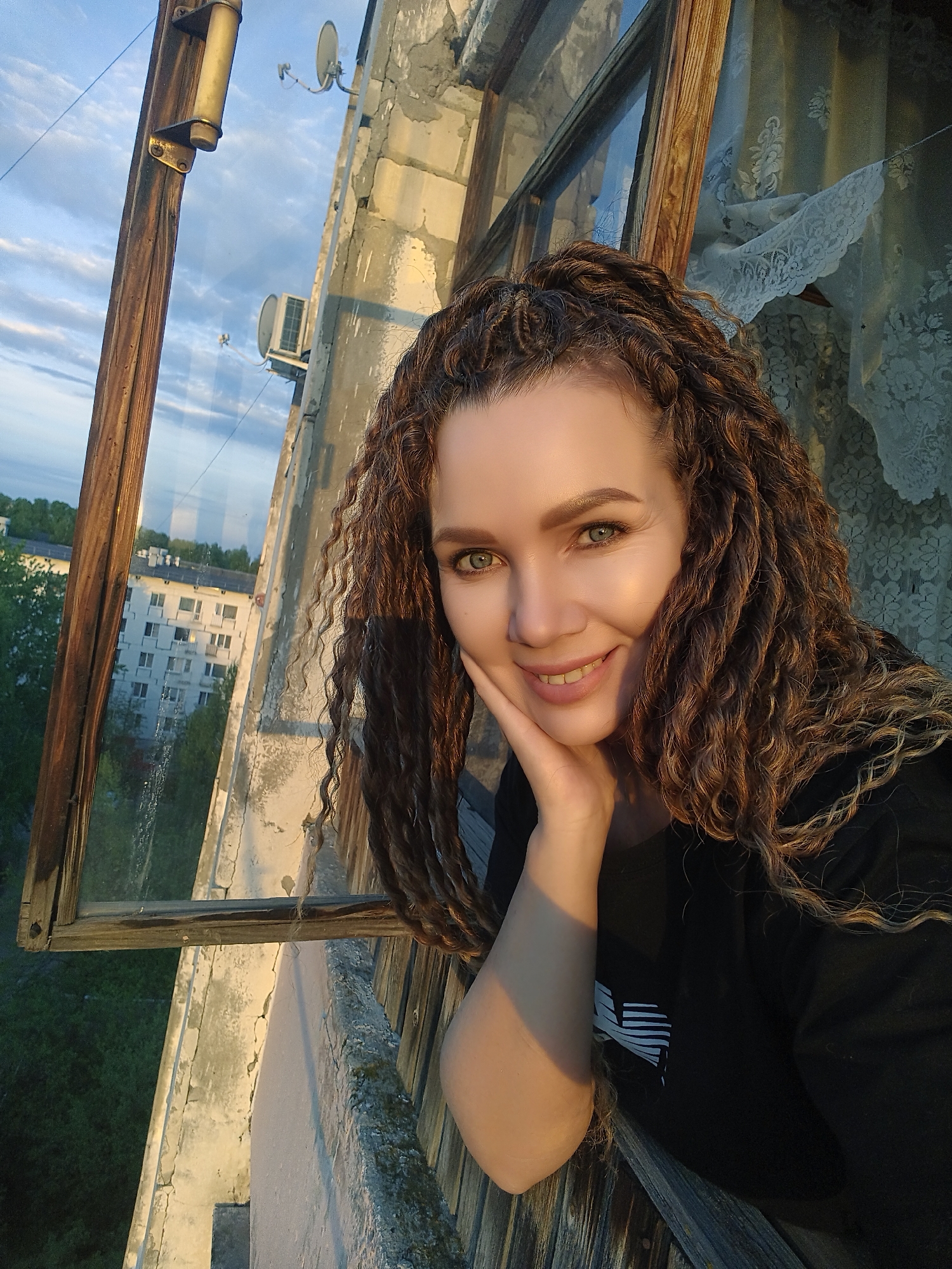 A medium shot captures a woman positioned near an open window in what appears to be a residential building. The woman is smiling, with her face resting on her hand. Her hair is curly and long, and she is wearing a dark-colored top.
Through the window, a view of a cityscape is visible. There are buildings and trees, suggesting an urban or suburban environment. The time of day appears to be late afternoon or early evening, judging by the angle and color of the sunlight.
The window frame is wooden and seems aged, hinting at the building's possible age. The surroundings visible through the window suggest that the location is in a city. The lace curtains in the background near the window.