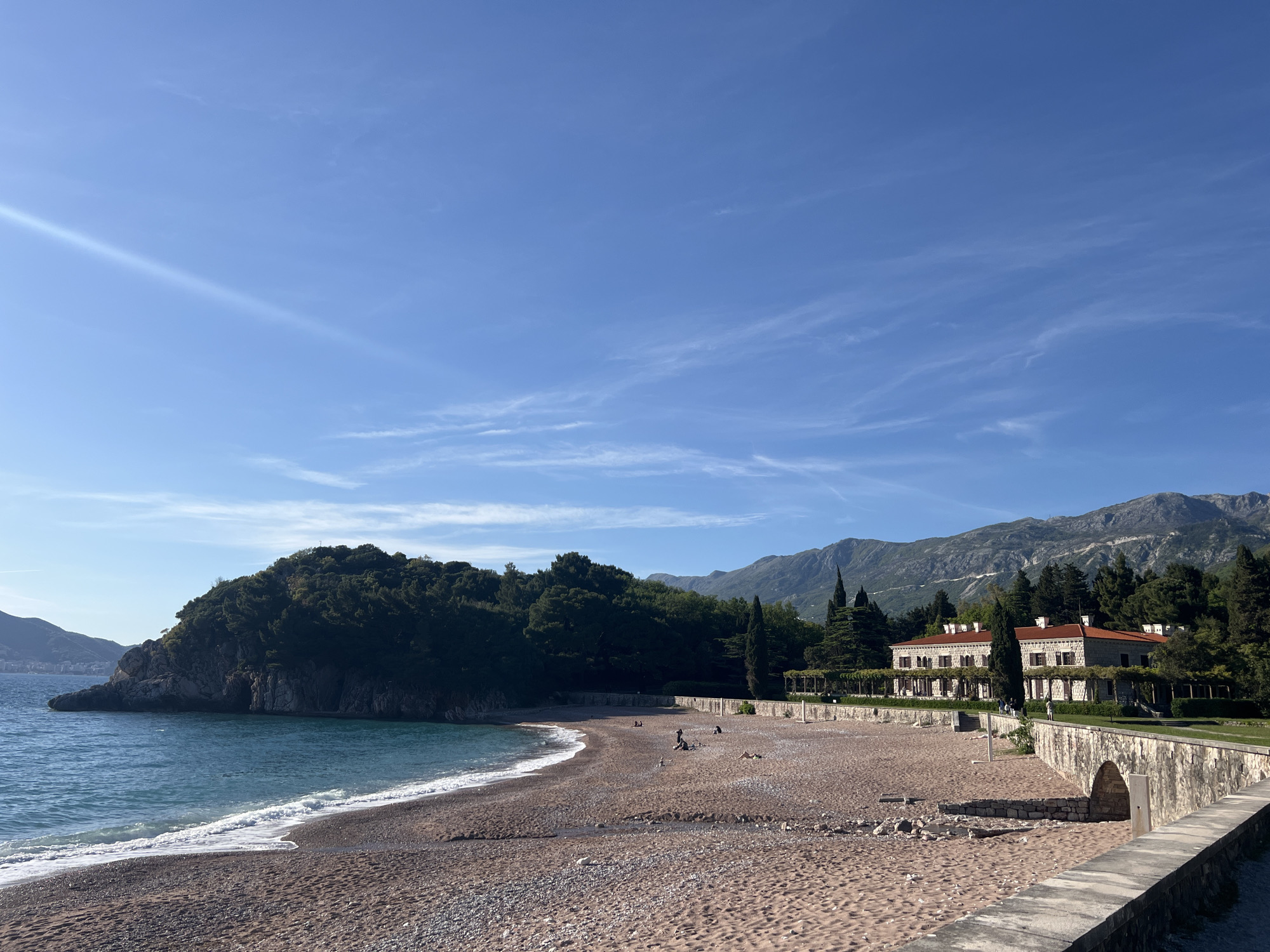 The image presents a serene coastal landscape in Sveti Stefan, Montenegro, under a bright blue sky dotted with wispy clouds. In the foreground and mid-ground, a crescent-shaped beach of sand and small pebbles meets the gentle, turquoise waves of the sea. To the left, a prominent, heavily forested headland extends into the water.
Along the right side of the beach, a grand, elegant building with red-tiled roofs, light-colored stone walls, and numerous windows with dark shutters stands nestled among tall green trees, some appearing to be cypresses. A covered walkway or pergola structure extends from the building towards the beach. A robust stone wall or promenade, featuring an arched opening, borders the beach, separating it from the building's manicured grounds.
Several people are visible on the beach, scattered casually. They appear to be relaxing, with some individuals sitting on the sand and others possibly strolling. In the background, majestic mountains rise, completing the scenic view.
The scene suggests a clear and pleasant day, likely mid-morning or late afternoon, given the bright sunlight and the shadows cast by the trees and building. The overall atmosphere is tranquil and inviting, showcasing the natural beauty and distinctive architecture of the location. No discernible text is visible in the image.