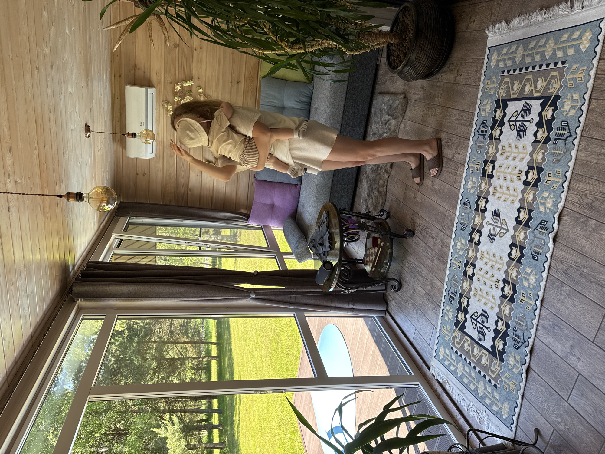 This image captures an indoor-outdoor living space in Копнино, Russia, characterized by extensive natural light and a rustic aesthetic. The scene features one adult female holding a young child, standing on a wooden floor in front of a grey sofa.
The adult female, with long light hair, wears a light-colored t-shirt, matching shorts, and brown slides. She holds a young child, likely a toddler, dressed in a light-colored bucket hat, t-shirt, and striped shorts. The adult's left hand is near the child's hat. The child is facing and embracing the adult.
The interior space features light-colored wood paneling on the walls and ceiling. Two pendant lights with large, exposed Edison-style bulbs hang from the ceiling. A white wall-mounted air conditioning unit is positioned on the wood-paneled wall. Below the air conditioner, a metallic wall decoration resembling golden leaves or butterflies is affixed.
Along the left and back walls, large floor-to-ceiling glass windows and doors offer a clear view of a lush green lawn and numerous trees under bright sunlight. Grey curtains with tie-backs are drawn to the side of the windows. Outside, a wooden deck is visible, with a light blue circular object (possibly a pool cover or small pool) placed on it.
To the left of the sofa, an ornate, two-tiered metal and glass side table is present. On its top tier, a black portable speaker and a folded dark garment are placed. The bottom tier holds a small wooden chess set box and a red box. A small grey faux-fur rug is positioned beneath the side table and partially under the sofa.
A patterned rug, primarily white with blue, grey, and yellow geometric and stylized tree designs, is centrally placed on the wooden floor. To the right of the sofa, a large potted plant with abundant green foliage stands in a dark, textured pot. Part of another plant is visible in a similar pot in the bottom left foreground.